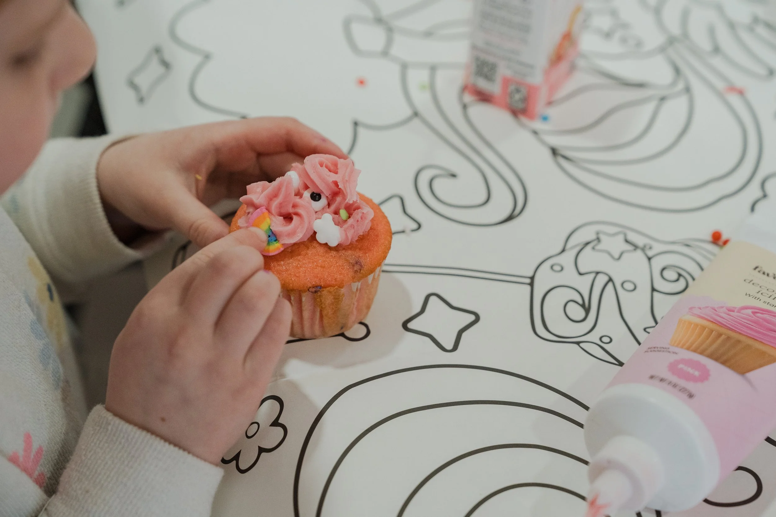 Picture of the hands of a child holding a cupcake while decorating it with pink frosting and sprinkles in the shapes of rainbows, eyes and clouds.