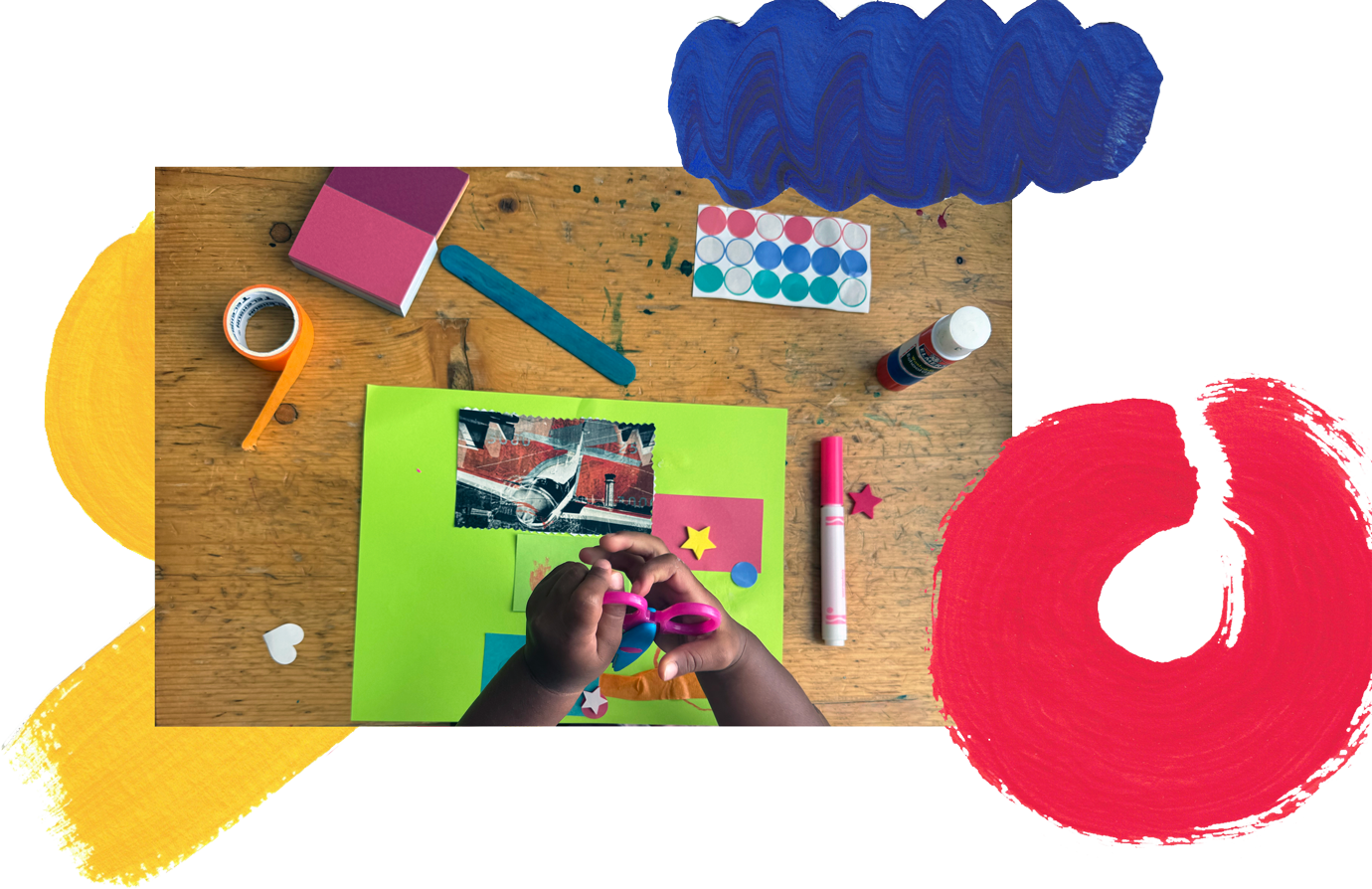 Child's hands working on crafting project with pink scissors, surrounded by colorful craft supplies on a wooden table, including glue, paint, paper, and decorative stars.