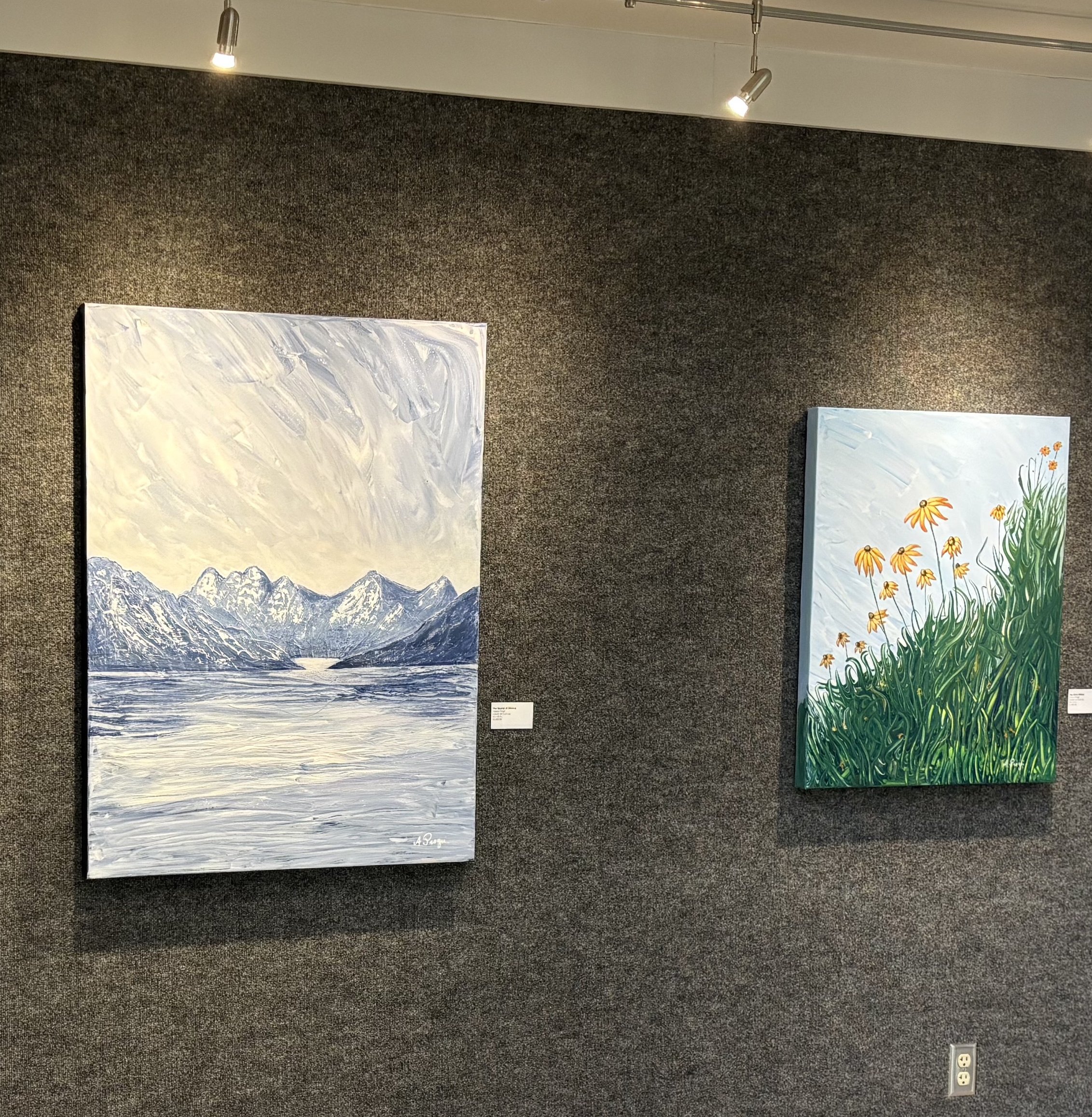Two paintings on a gallery wall; the left painting features a snowy mountain landscape with a blue sky, and the right painting depicts yellow flowers among tall green grass against a blue sky.