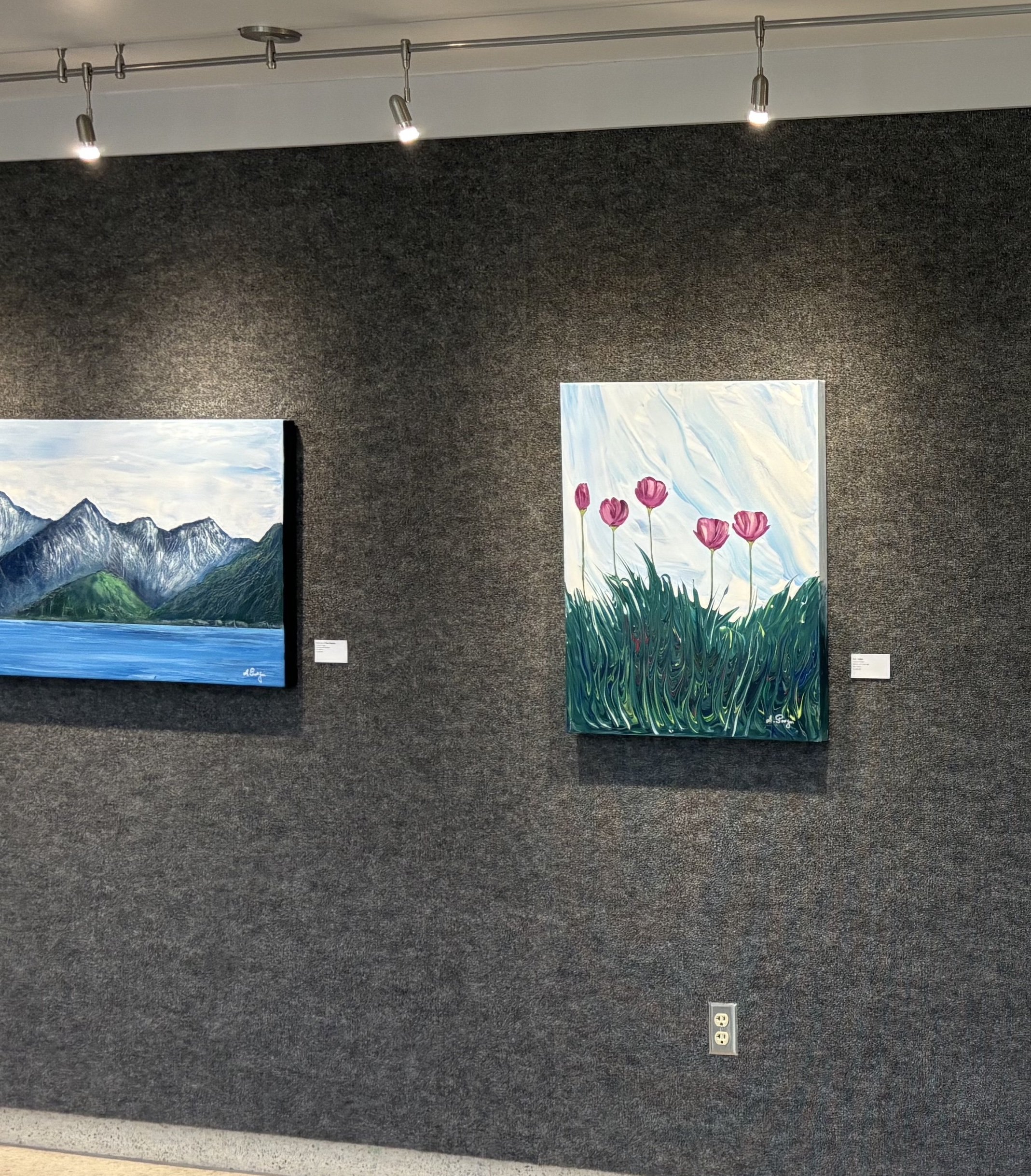 Art gallery with two paintings; one of mountains by a lake and the other of pink flowers in tall grass with a blue sky background.