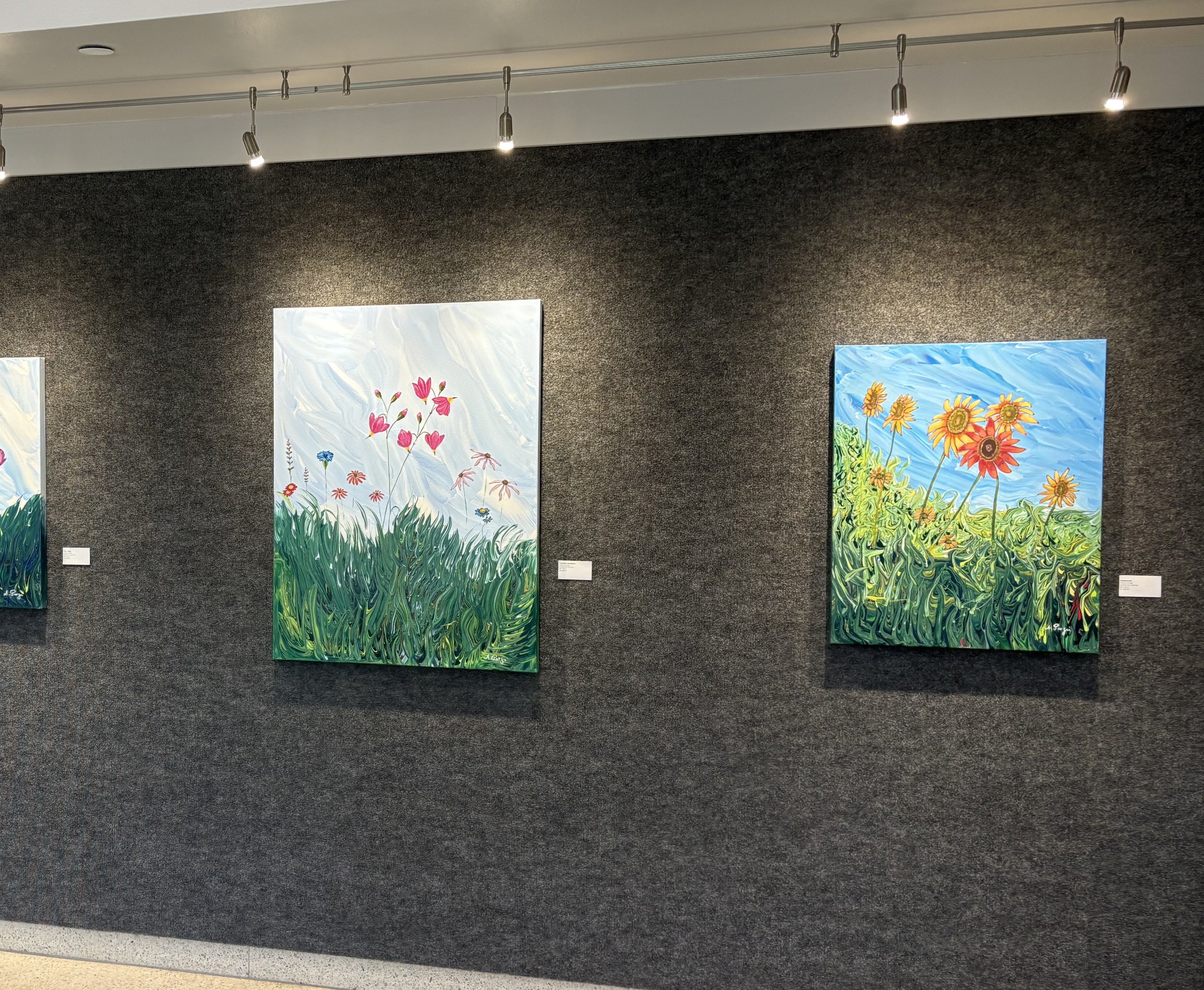 Gallery wall displaying two colorful paintings of flowers, each set against a backdrop of swirling blue skies and green foliage.