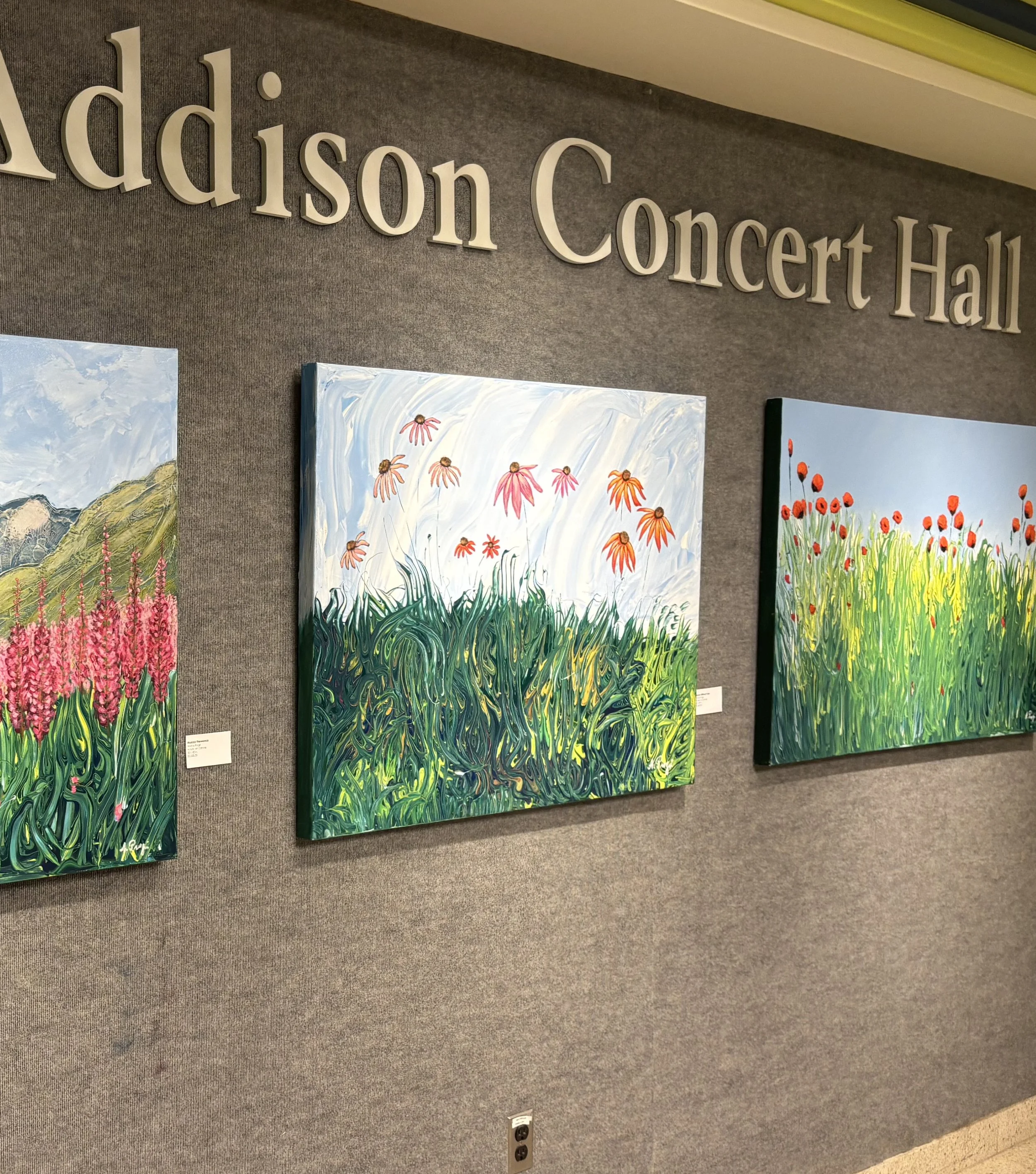 Art exhibit at Addison Concert Hall featuring three colorful floral paintings on a gray wall.