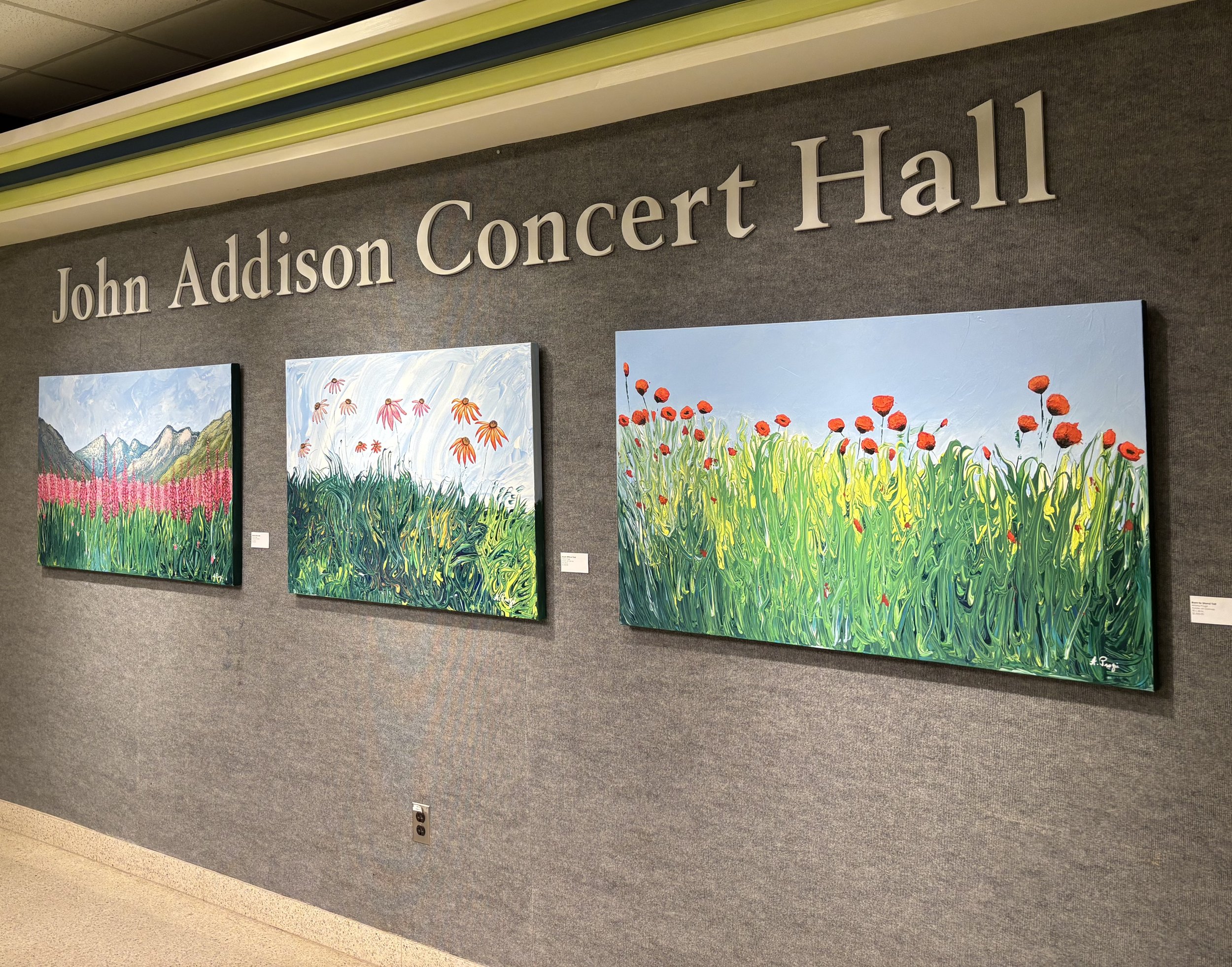 Wall display at John Addison Concert Hall featuring three colorful landscape paintings with fields and flowers.