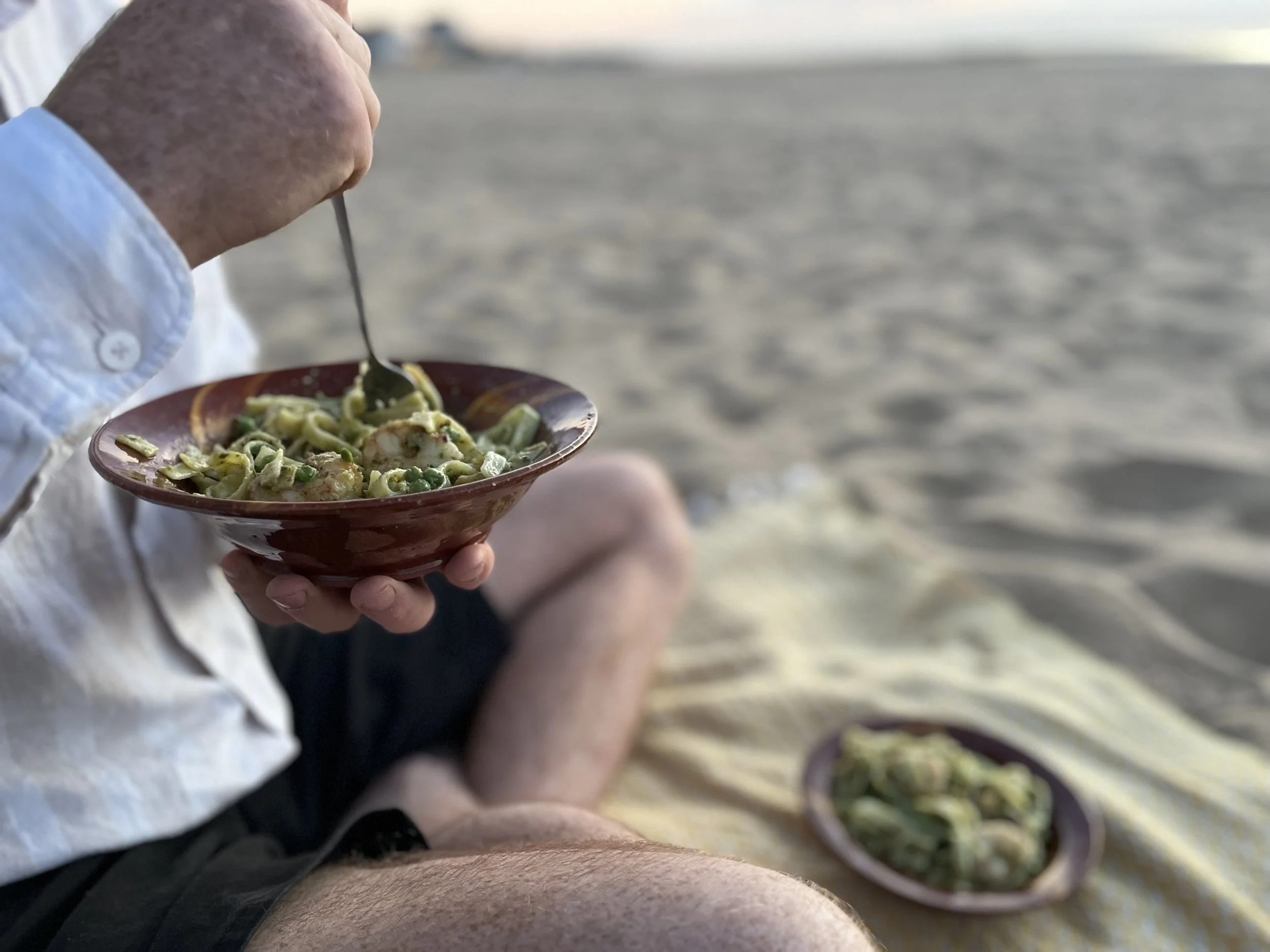 Person holding a bowl of pasta with green sauce, sitting outdoors near a water body.