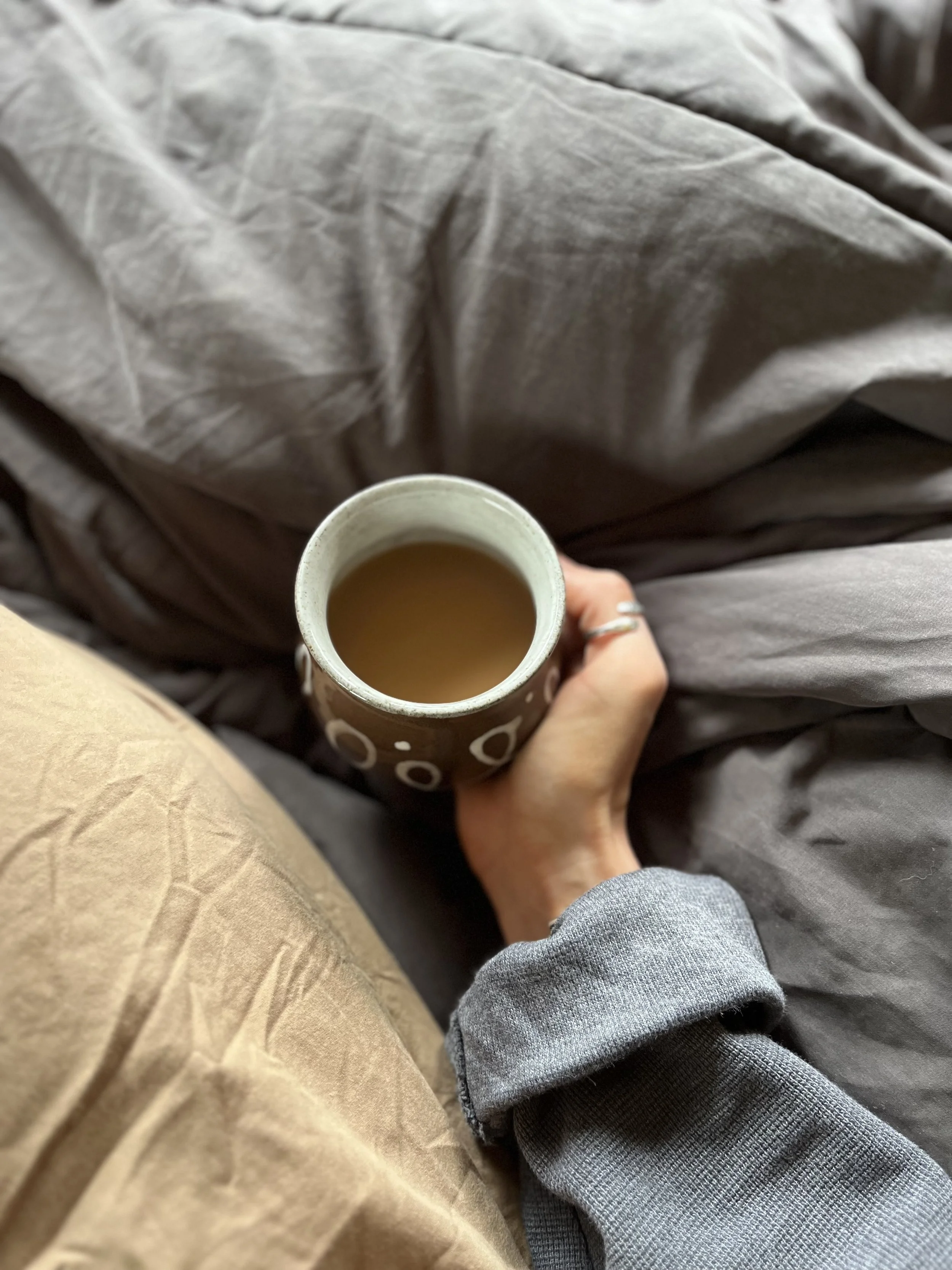 A person holding a coffee mug while sitting on a bed or sofa. The person is wearing a gray sweatshirt and beige pants, with a gray blanket or sheet nearby.