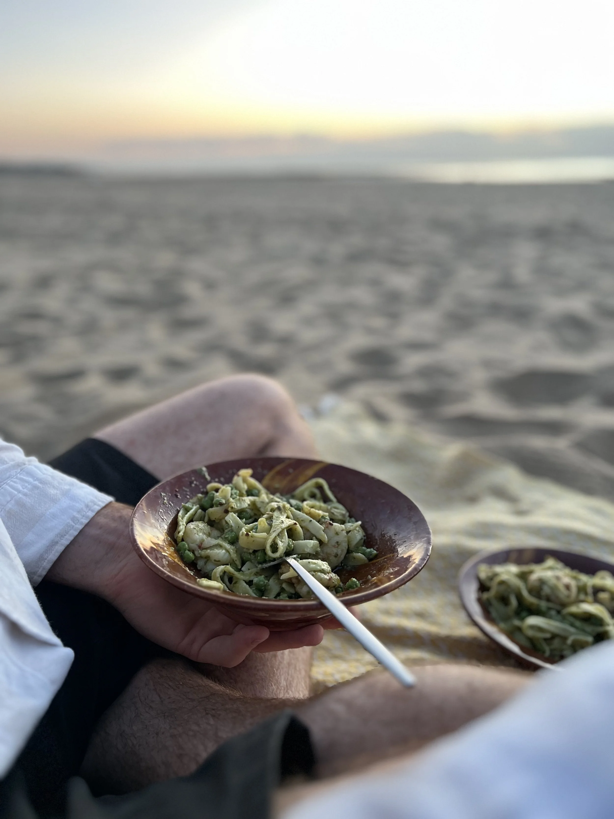 Person holding a bowl of pasta with peas at the beach during sunset.