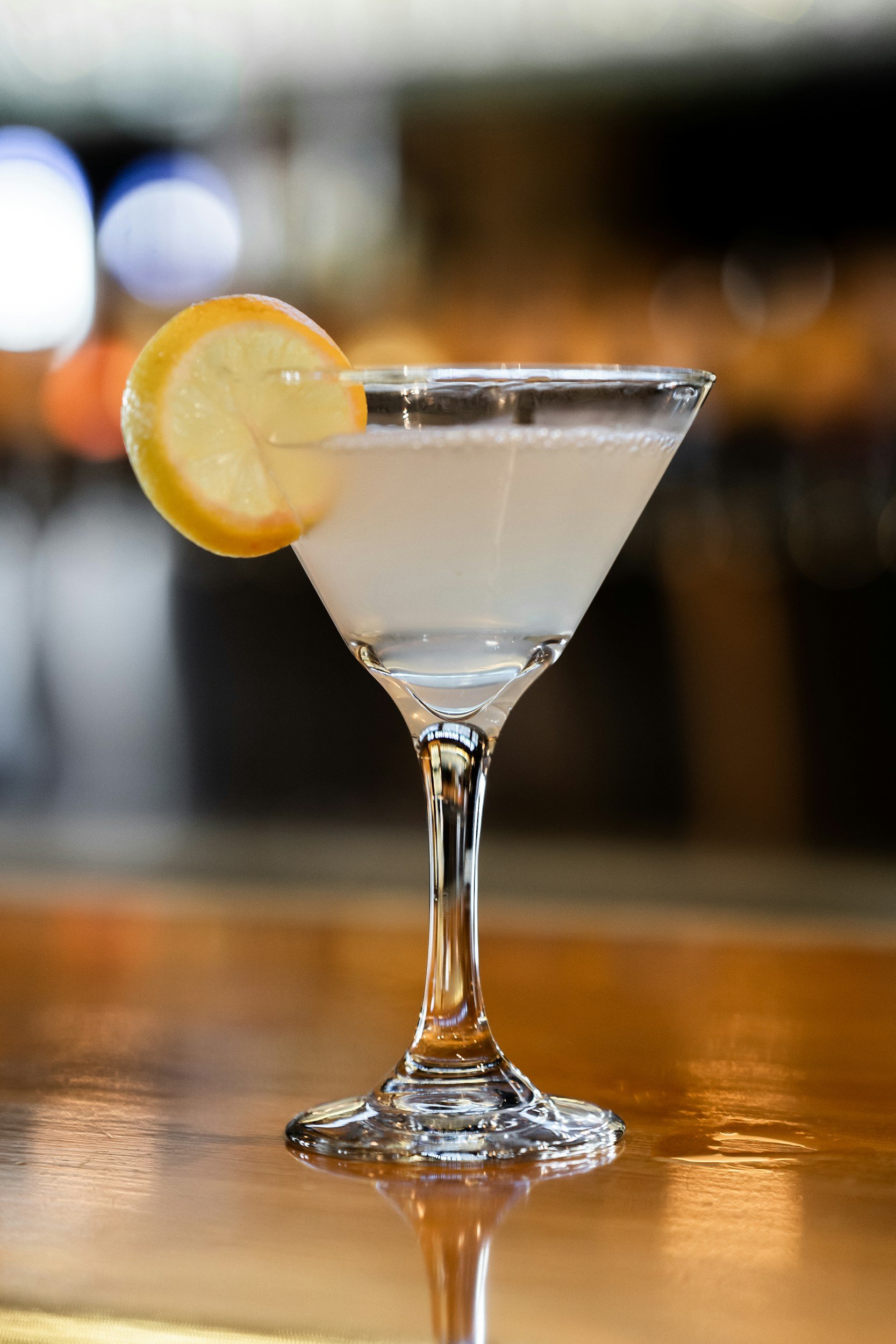 Martini glass with clear cocktail and lemon slice garnish on a wooden bar top.