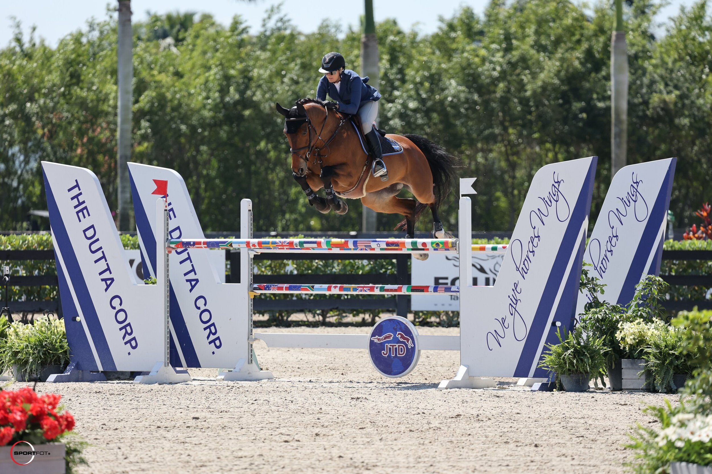 A rider and a horse jumping over a colorful obstacle in an equestrian event with sponsor banners and green trees in the background.