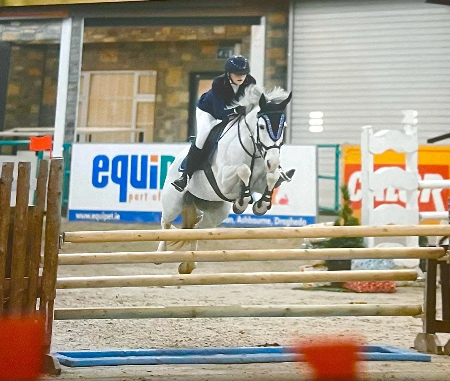A person riding a white horse jumping over a hurdle during an equestrian event inside an indoor arena.