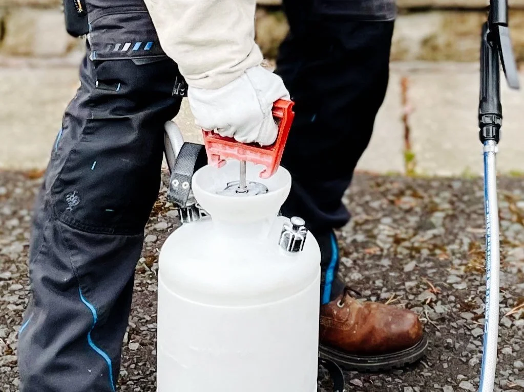 Person wearing black pants and brown boots, filling a large white container with a handheld pump sprayer, using a red tool. 