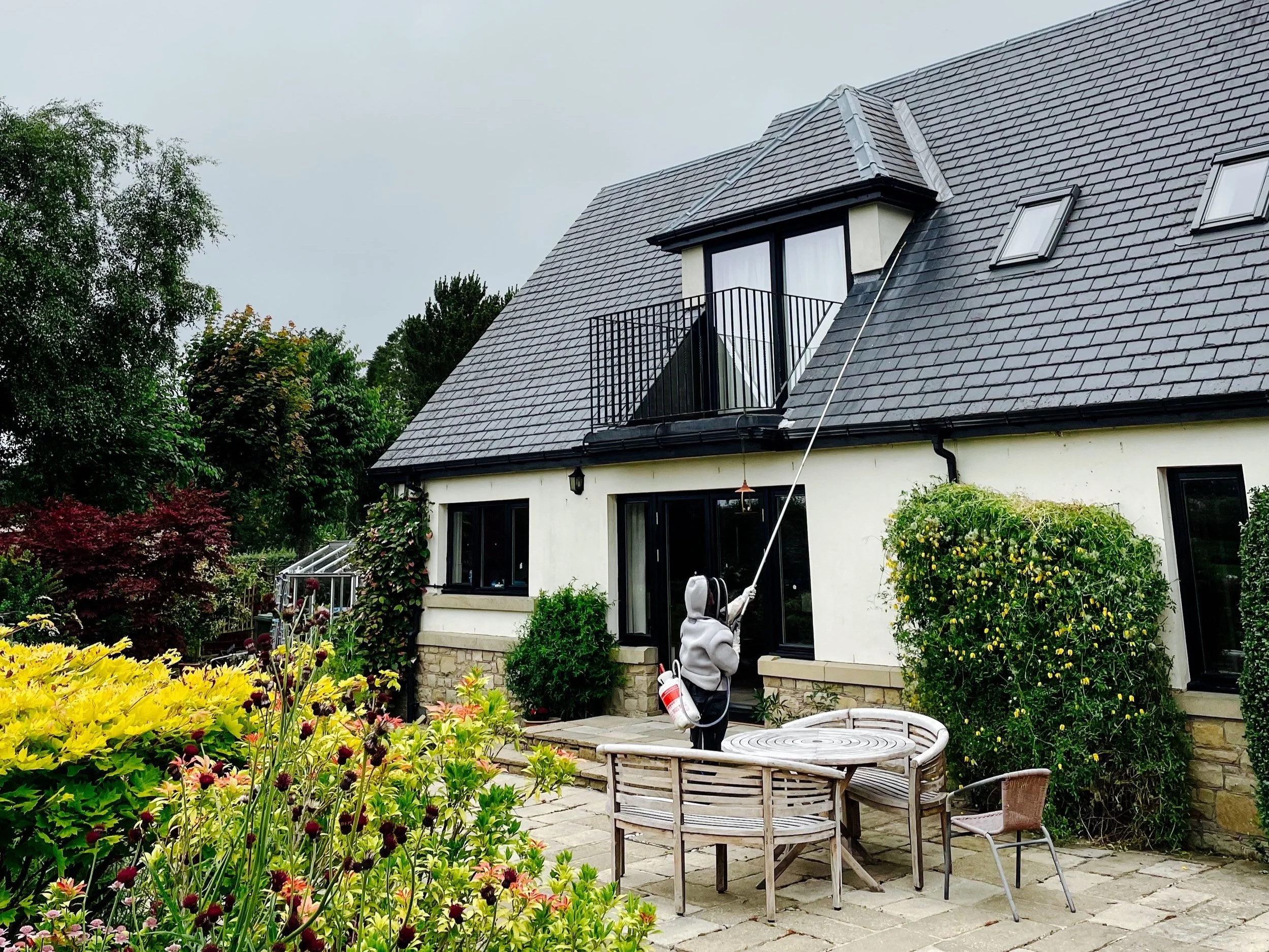A person in a bee suit spraying chemicals on a wasp nest with an extendable pole in a backyard garden with colourful flowers and shrubs.