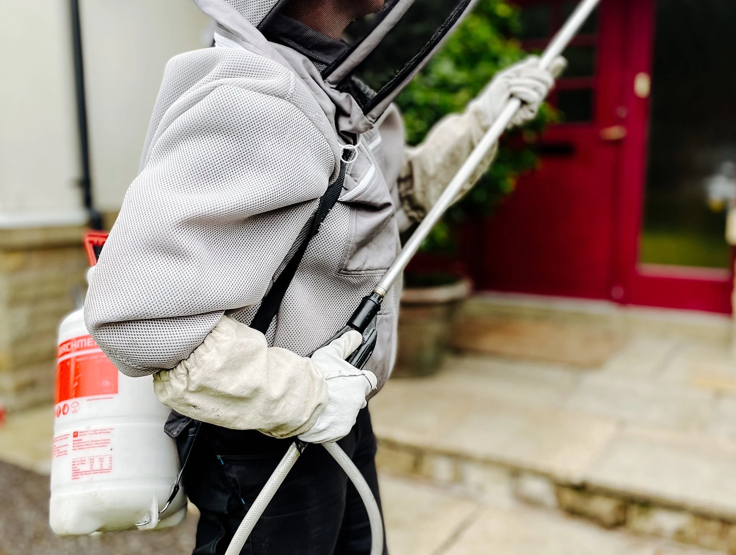 A person dressed in protective gardening gear, including gloves, holding a sprayer wand, and carrying a container of chemicals, standing in front of a building with a red door.