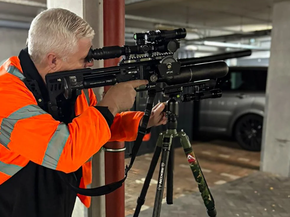 A man with gray hair wearing a bright orange safety jacket looking through a rifle scope mounted on a black rifle with a bipod, positioned in an indoor parking garage.
