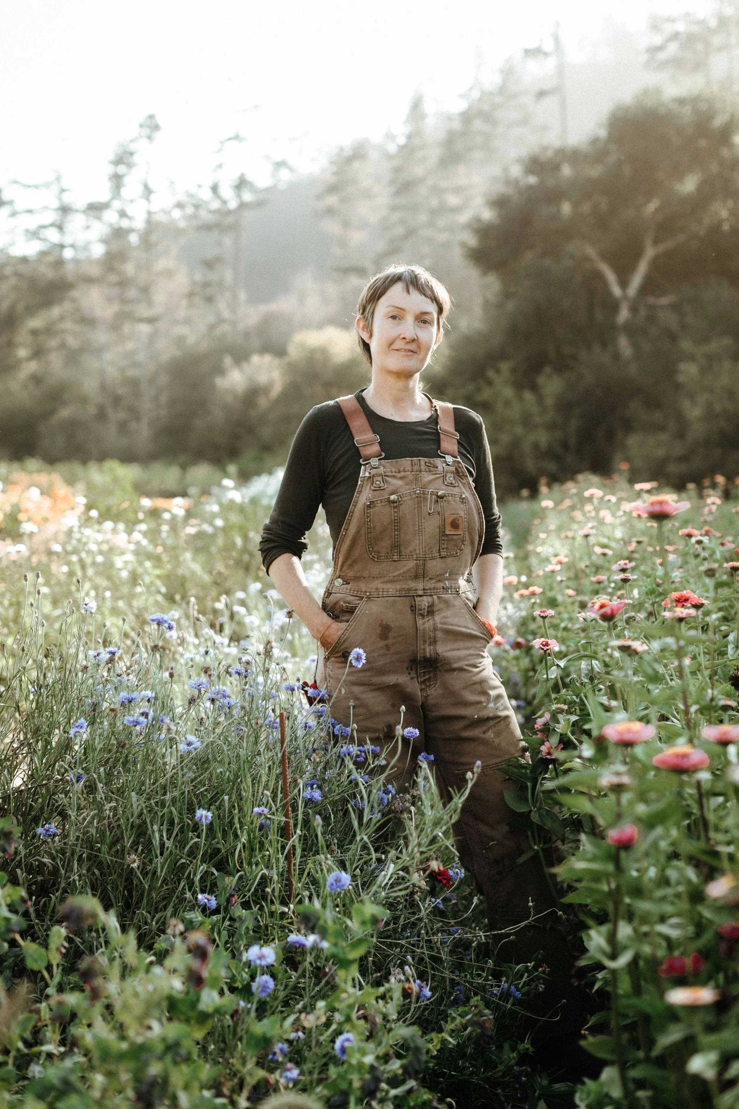 A woman standing in a flower field, wearing brown overalls and a black long sleeve shirt, with trees and a misty landscape in the background.