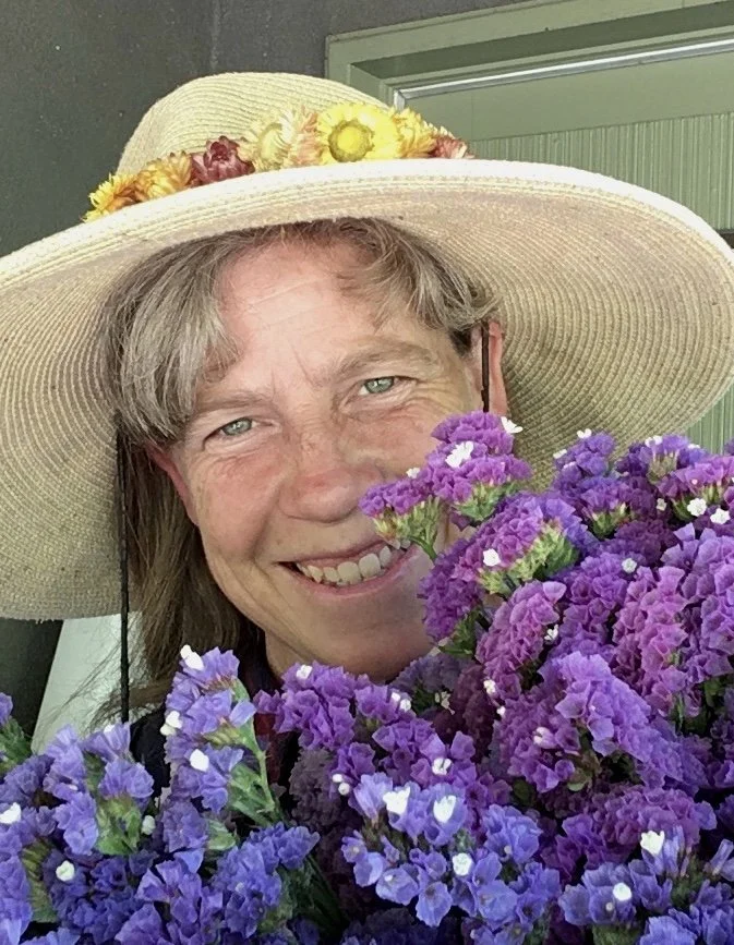 A woman with light brown hair and blue eyes wearing a large straw hat with a flower crown and holding a cluster of purple flowers.