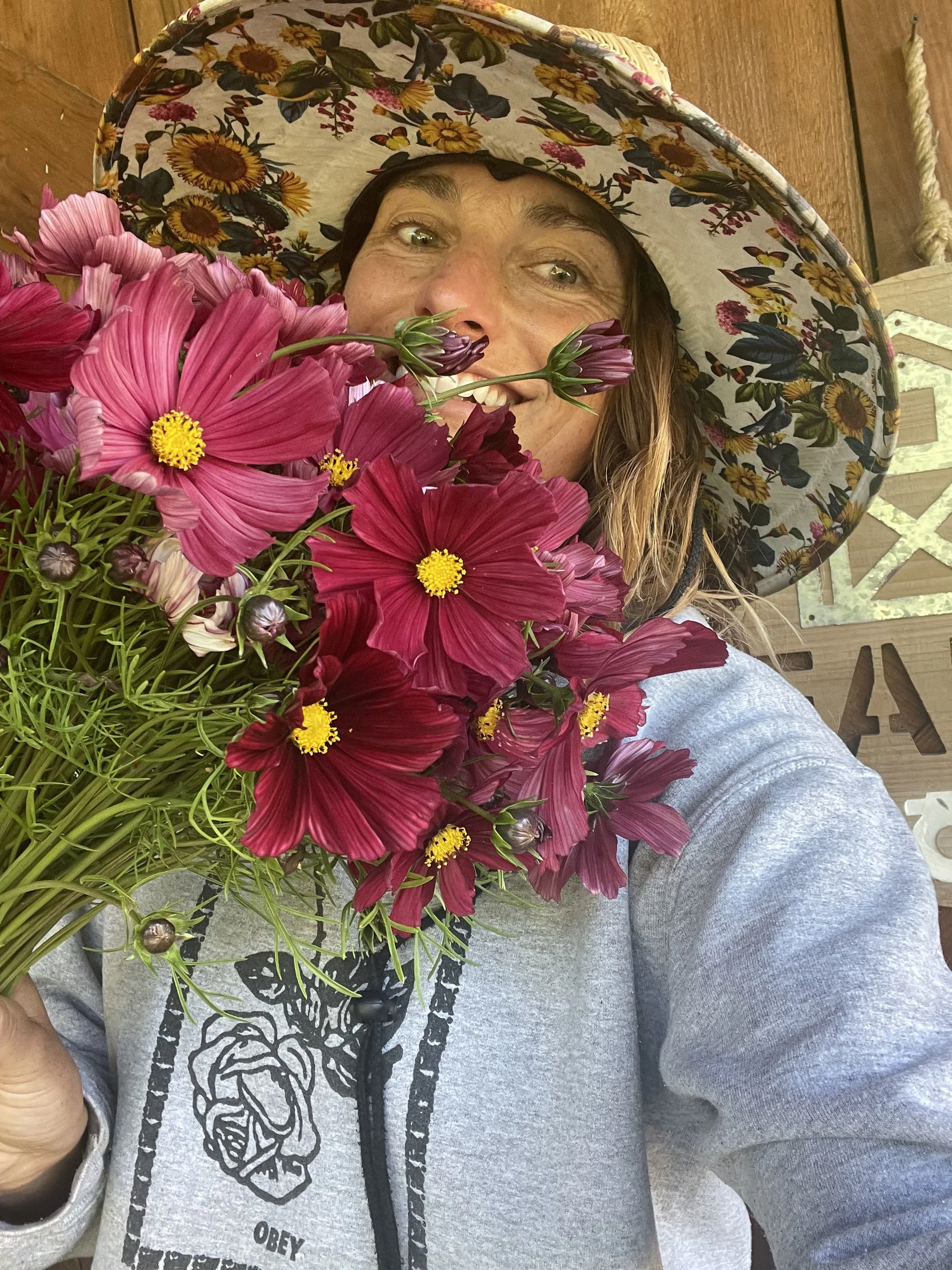 A woman taking a selfie holding a large bouquet of pink and deep red flowers, wearing a floral wide-brimmed hat and a gray hoodie with a rose and