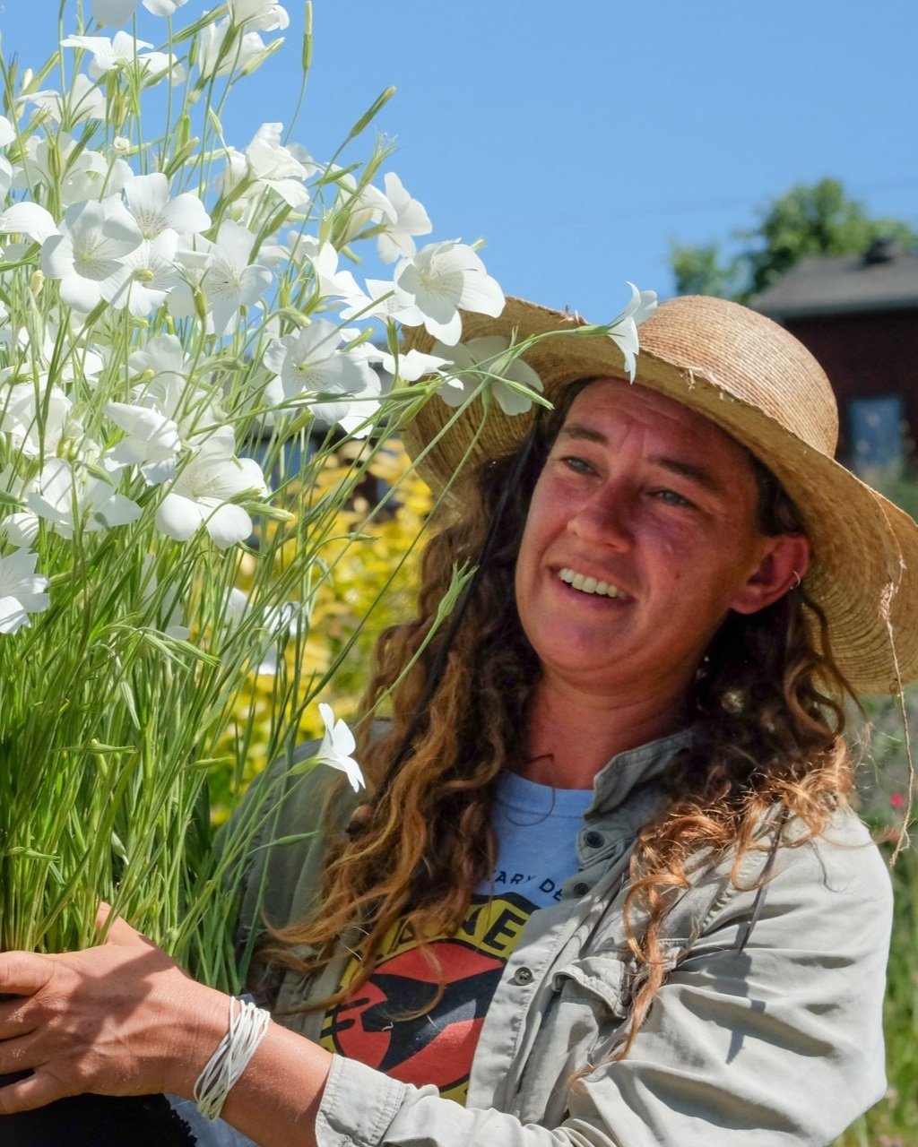 A woman with long curly hair wearing a wide-brim straw hat, a grey jacket, and a graphic t-shirt, holding a bouquet of white flowers outdoors on a sunny day.