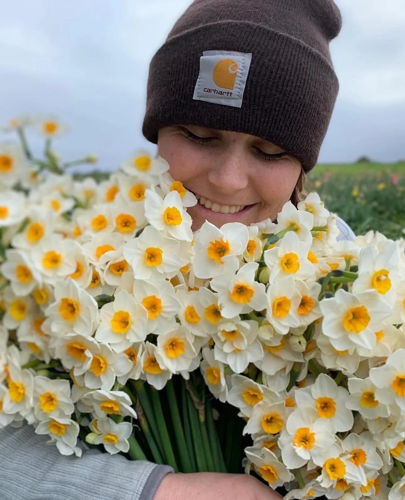 Person wearing a brown Carhartt beanie smiling and holding a large bouquet of white flowers with yellow centers in an outdoor field.