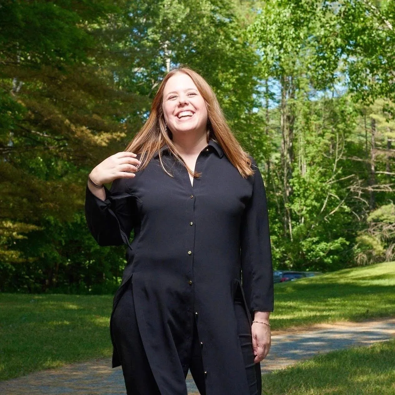 Smiling woman in black shirt standing outdoors in a green park with trees and grass.