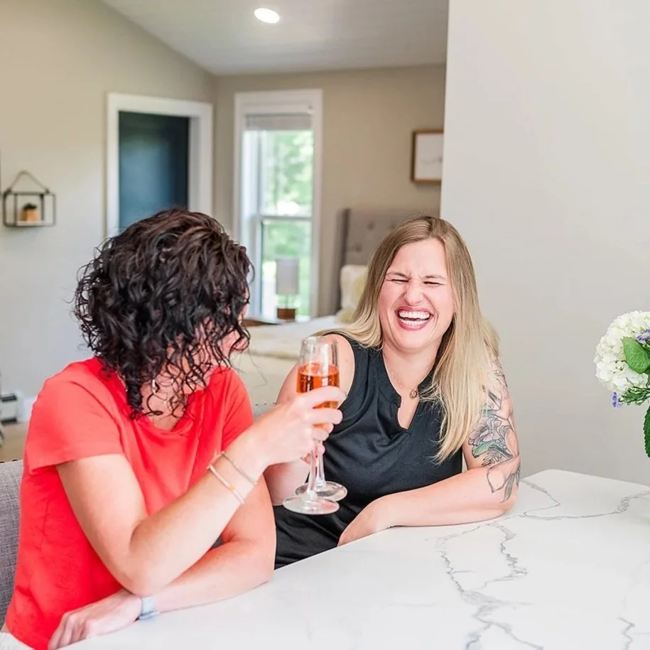 Two women sitting at a table enjoying drinks. One woman has curly dark hair and is wearing a red shirt, holding a wine glass. The other woman has straight blonde hair, is wearing a black shirt, and is laughing with her eyes closed.