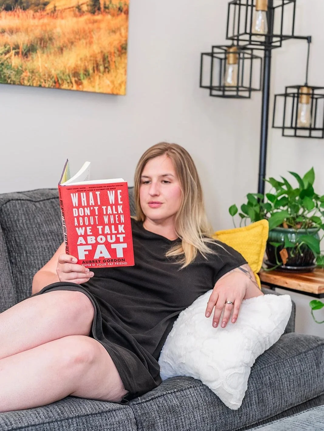 blonde woman in black dress sits on a couch and reads the book titled "what we don't talk about when we talk about fat"