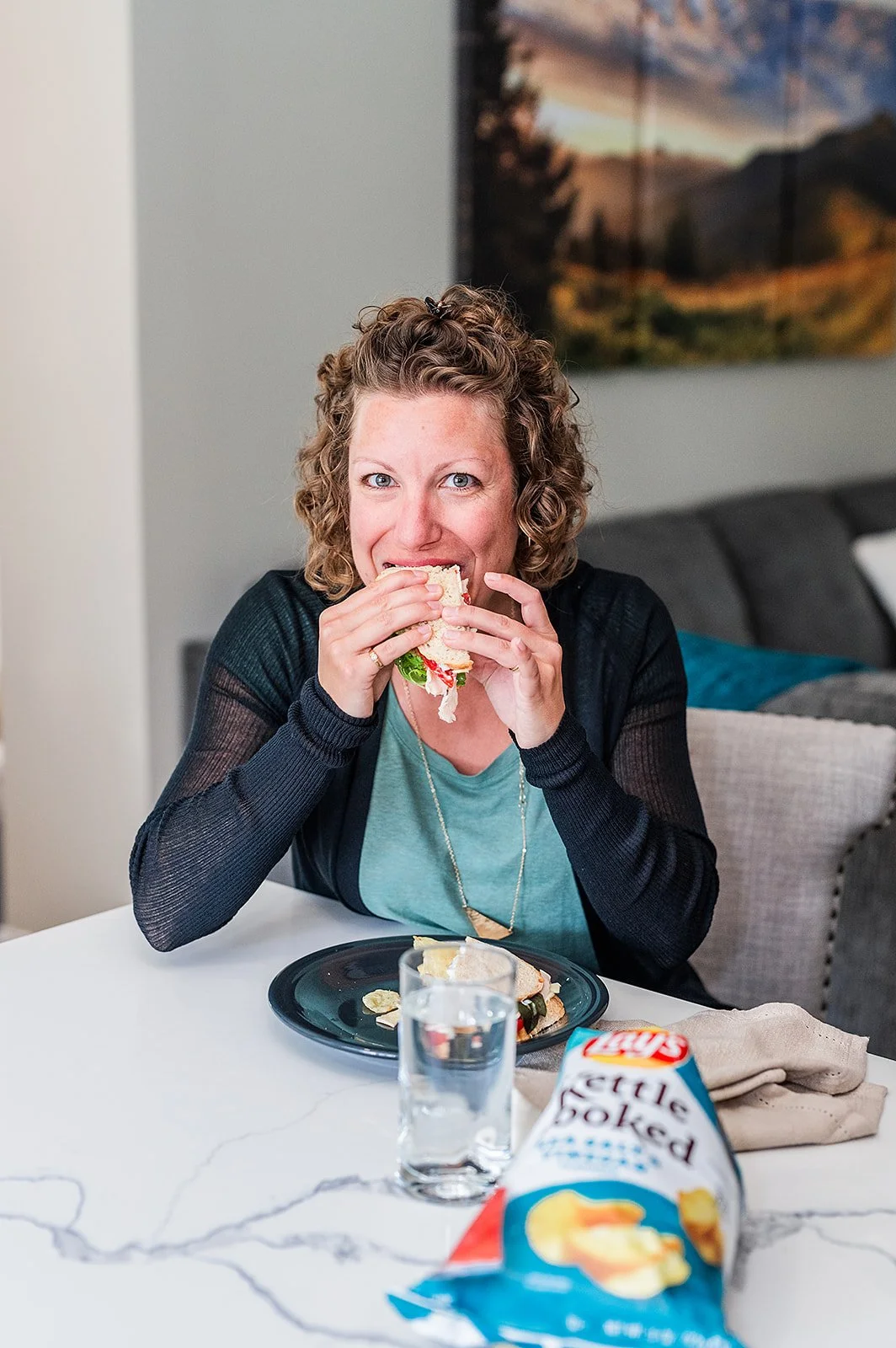 A woman with curly hair and a black top eating a sandwich at a white table. There is a glass of water, a bag of Lay's potato chips, and a beige napkin on the table, with a couch and landscape painting in the background.