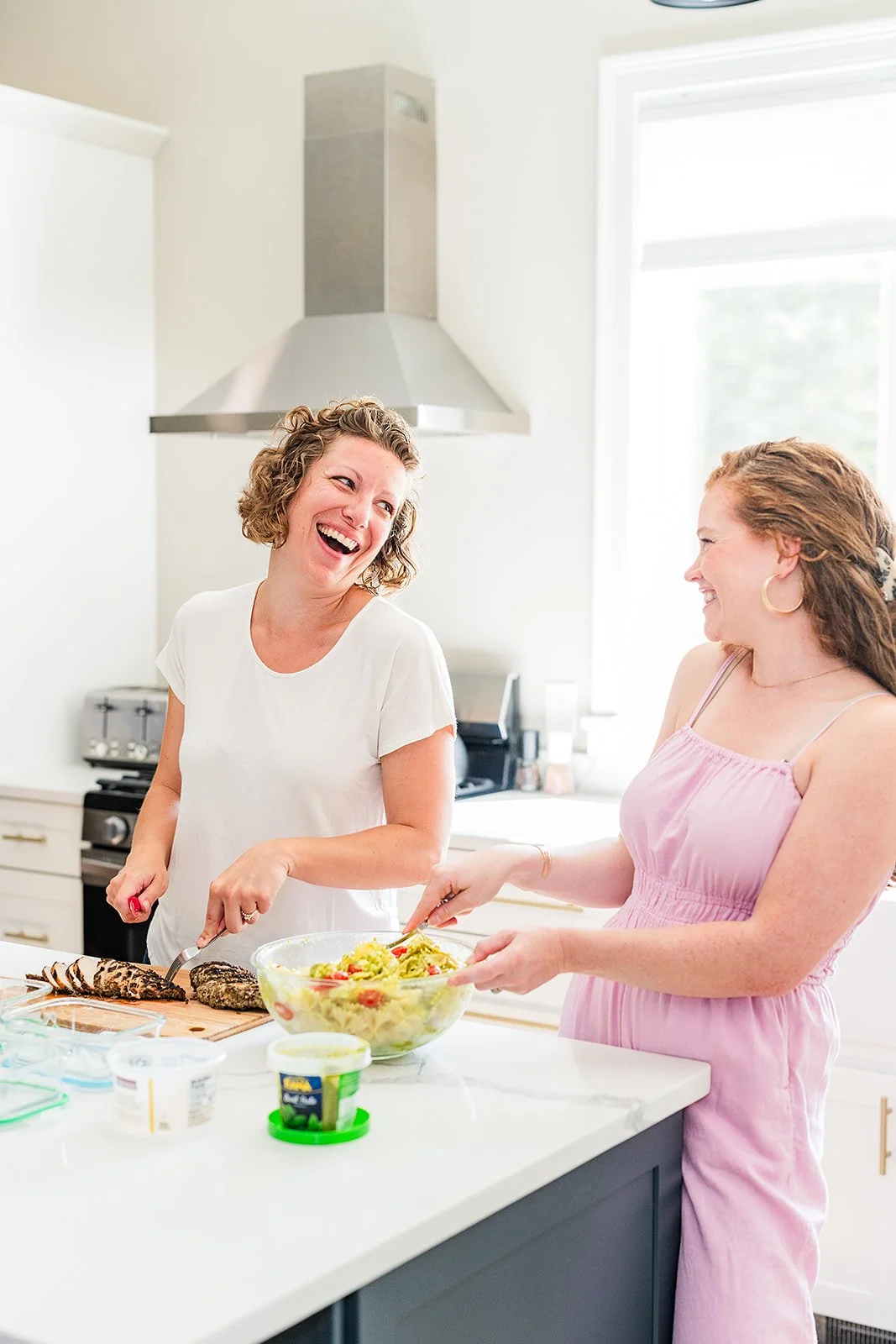 Two women laughing and enjoying each other's company while preparing food together in a bright kitchen. One woman is holding a bowl of guacamole and the other is pointing at it.