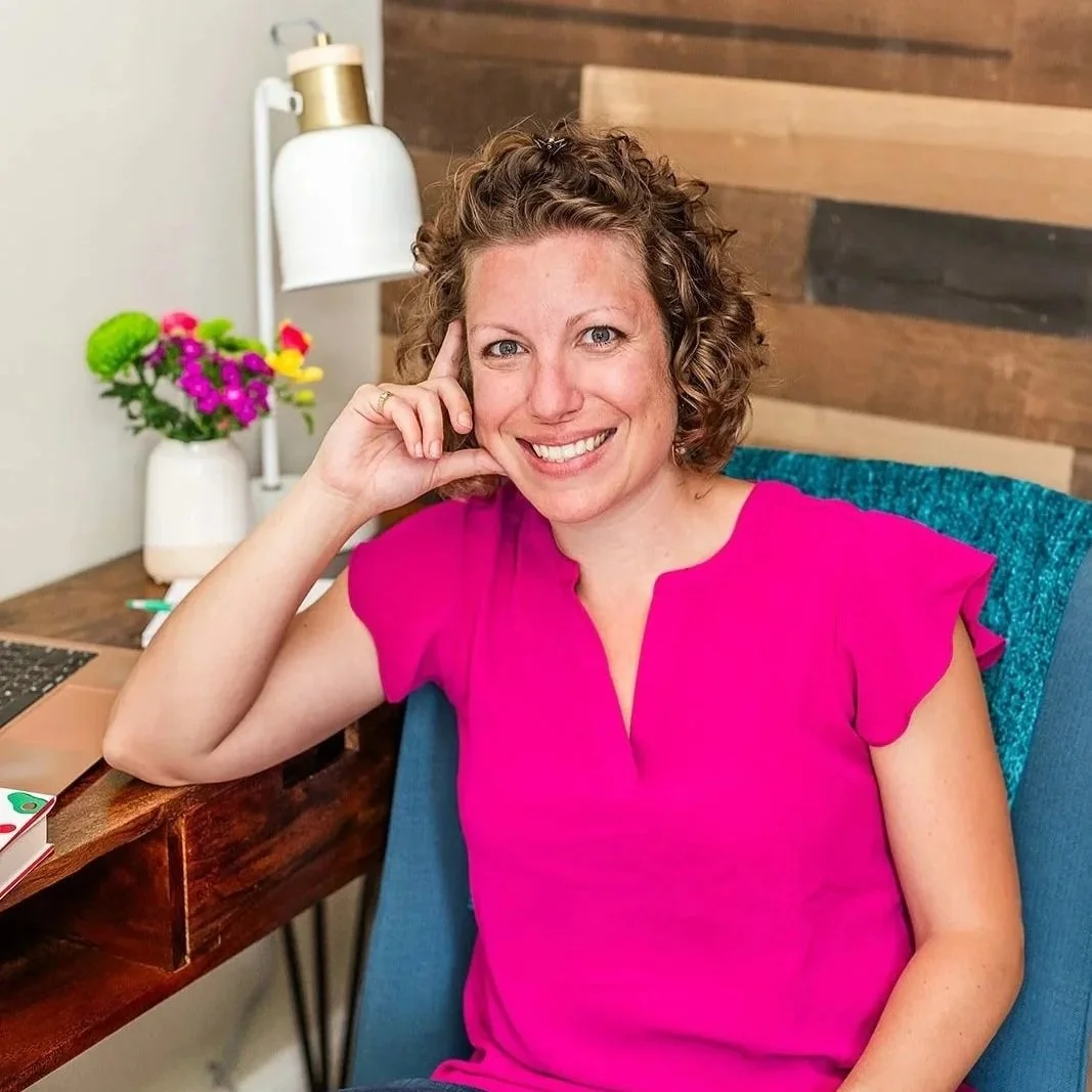 A woman with curly brown hair and a pink blouse sitting at a desk, smiling at the camera, with a lamp and a flower arrangement in the background.