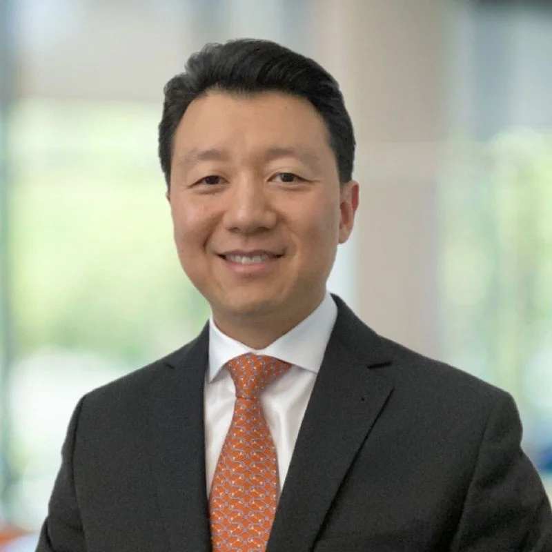 A professional man wearing a black suit, white shirt, and orange patterned tie, smiling in an office setting with blurred background.
