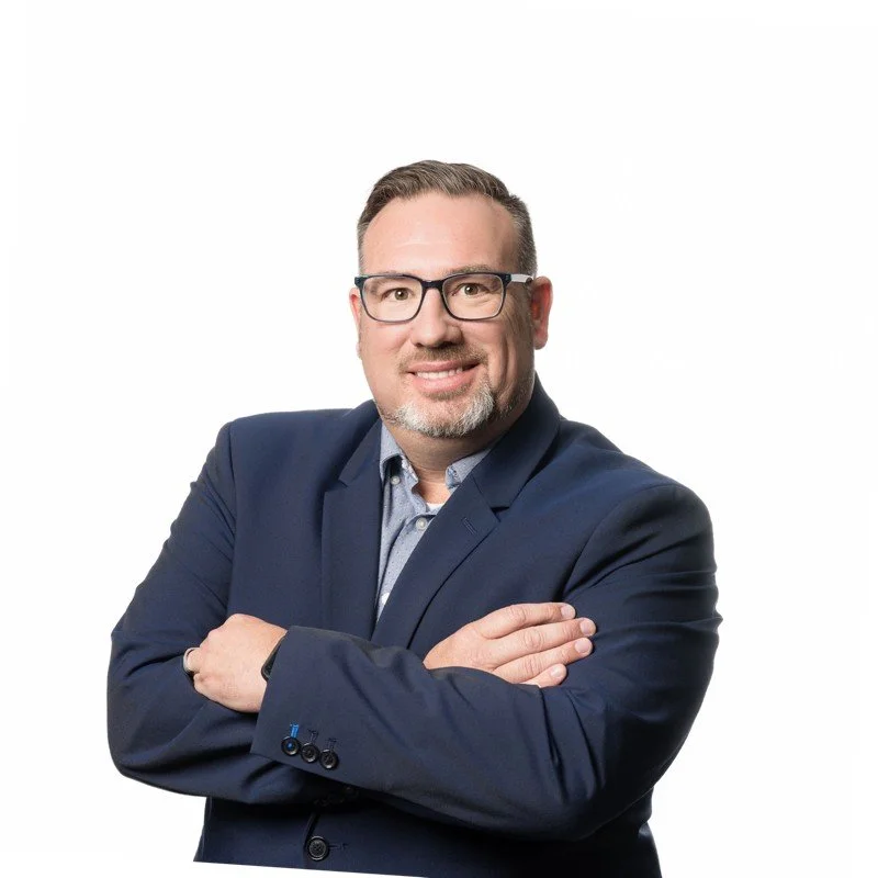A man with glasses, short brown hair, and a beard, dressed in a navy blazer and light blue shirt, smiling with arms crossed against a plain white background.