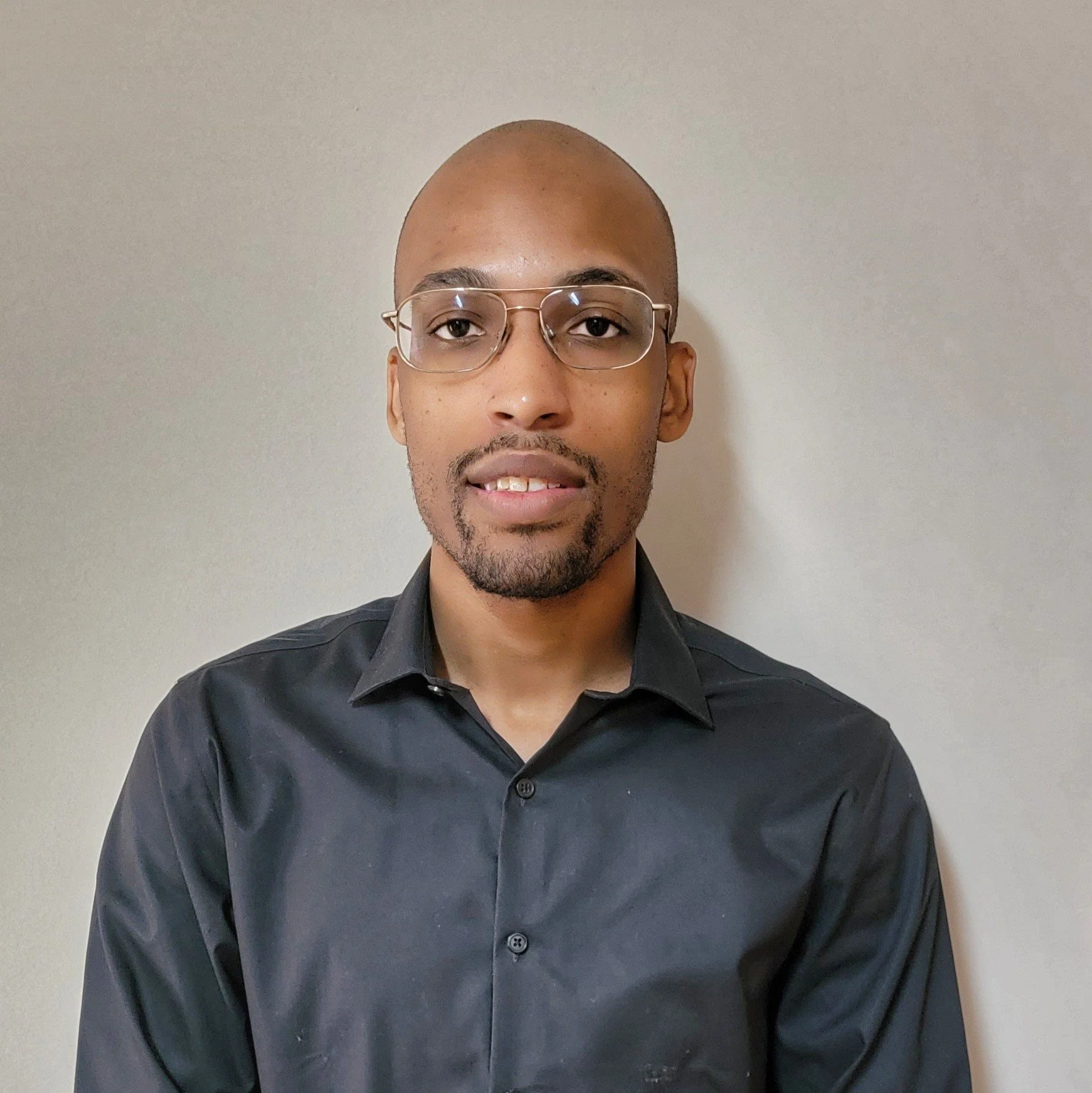 A man with a shaved head, glasses, and a short beard, wearing a black button-up shirt, stands against a plain light-colored wall.