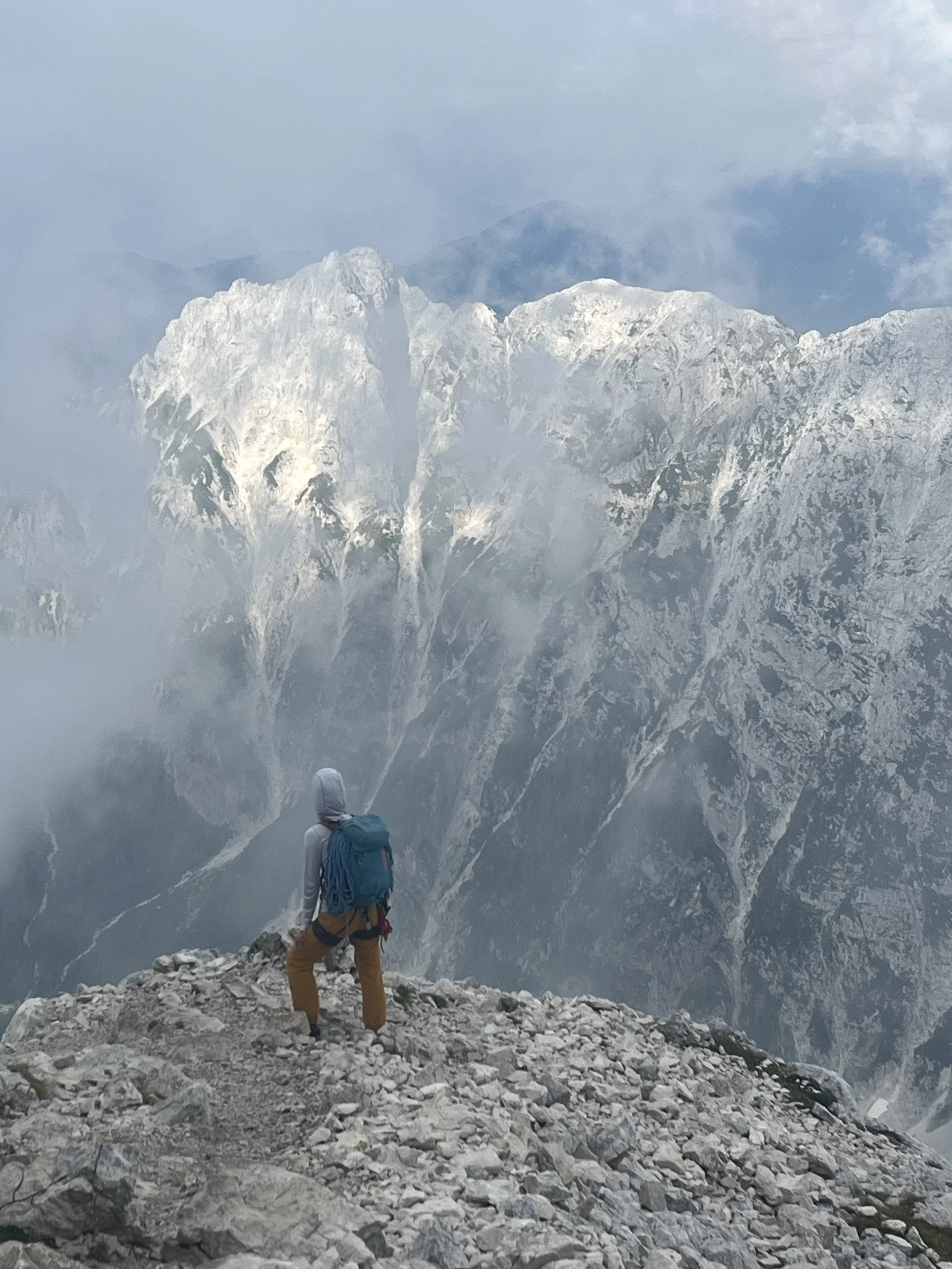 A person standing on rocky terrain overlooking snow-capped mountain peaks surrounded by clouds. Training for climbing big multipitch routes in Europe.