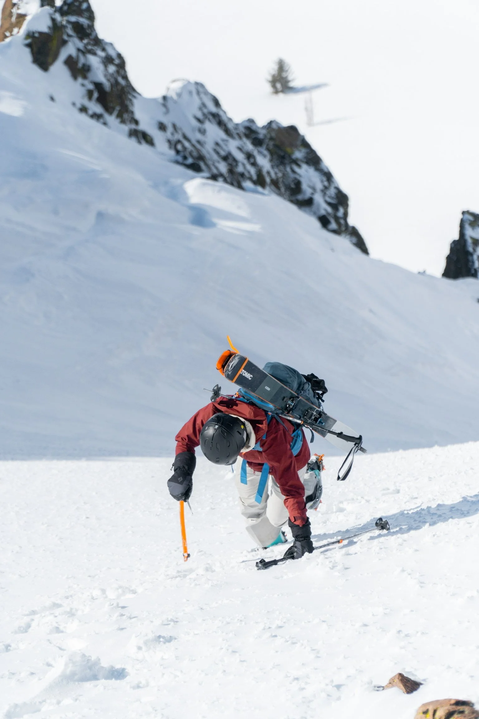 A person climbing snowy mountain terrain with ice axe, wearing helmet and winter gear, carrying equipment on their back, set against rocky cliffs and snow-covered landscape.