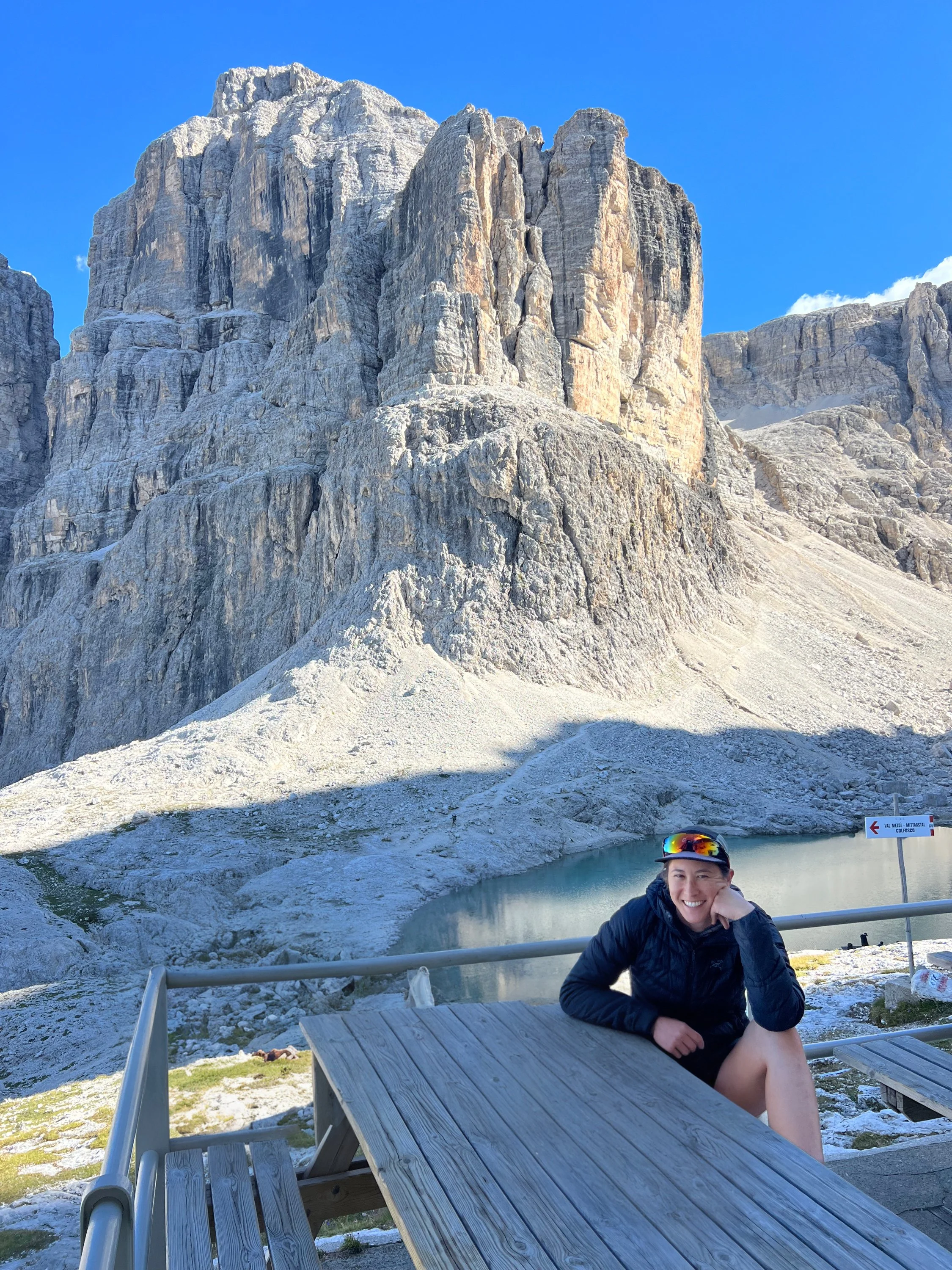 A woman smiling and sitting at a wooden picnic table outdoors with a mountain and a small lake in the background. Hut to hut running tour.
