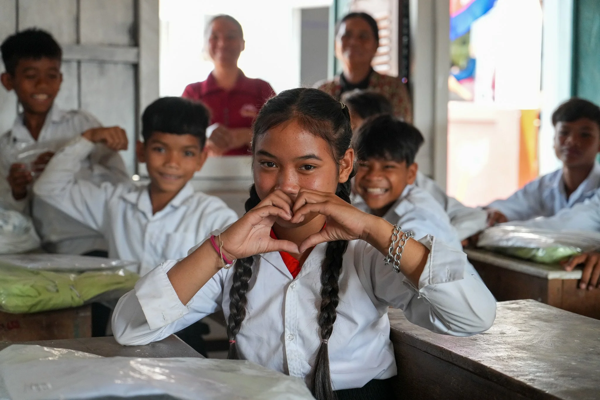 A young girl with long braided hair makes a heart shape with her hands while sitting at a desk in a classroom with other smiling students and two teachers in the background.