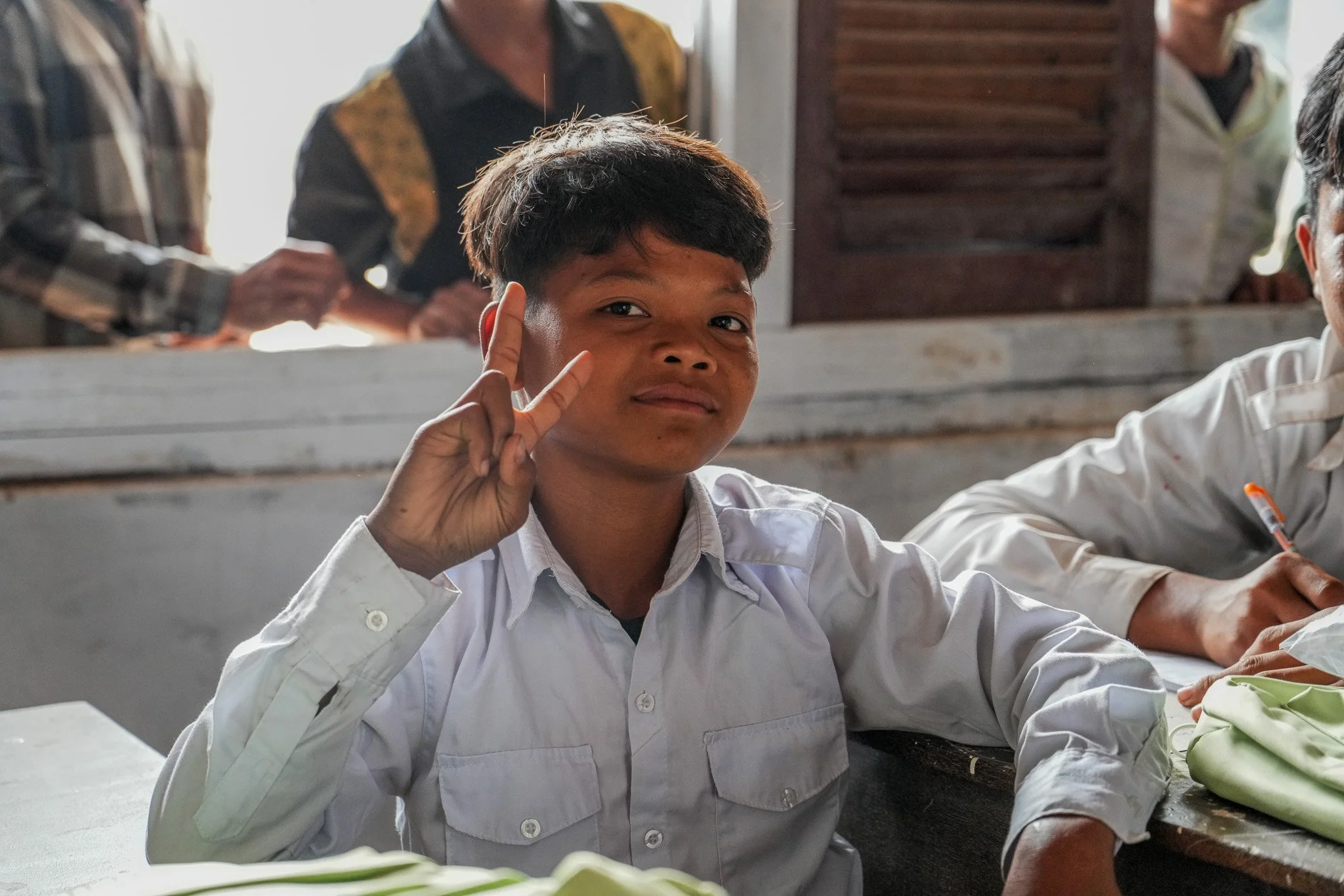 A young student in a white shirt making a peace sign with his right hand in a classroom setting.