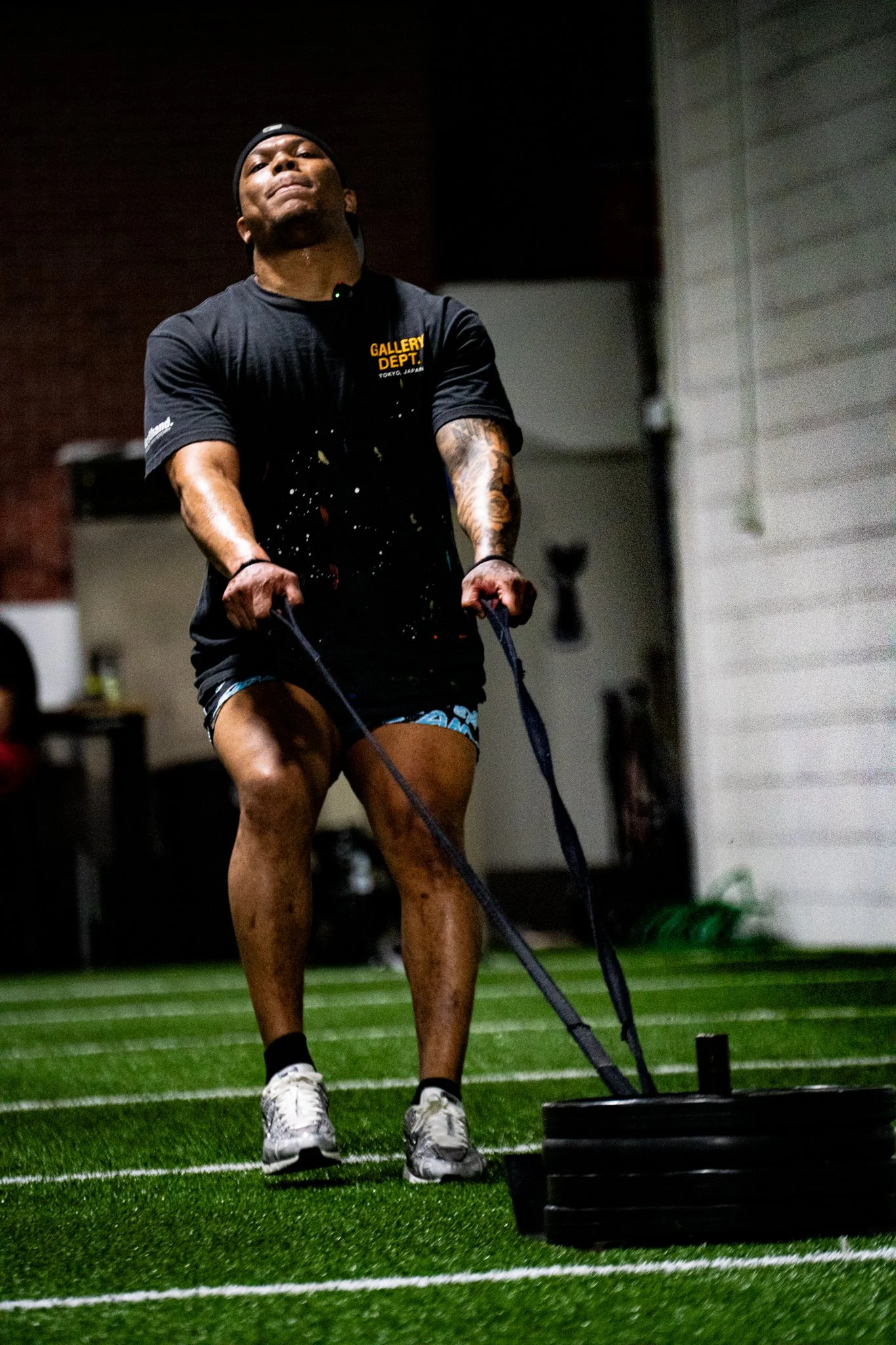 A man working out indoors on artificial turf, pushing a weighted sled. He is wearing a black T-shirt with yellow lettering, shorts, and athletic shoes.