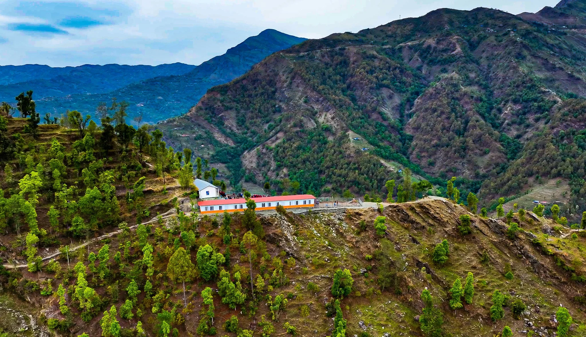 Mountain landscape with green trees, a white and orange building on a hillside, and rugged terrain in the background.