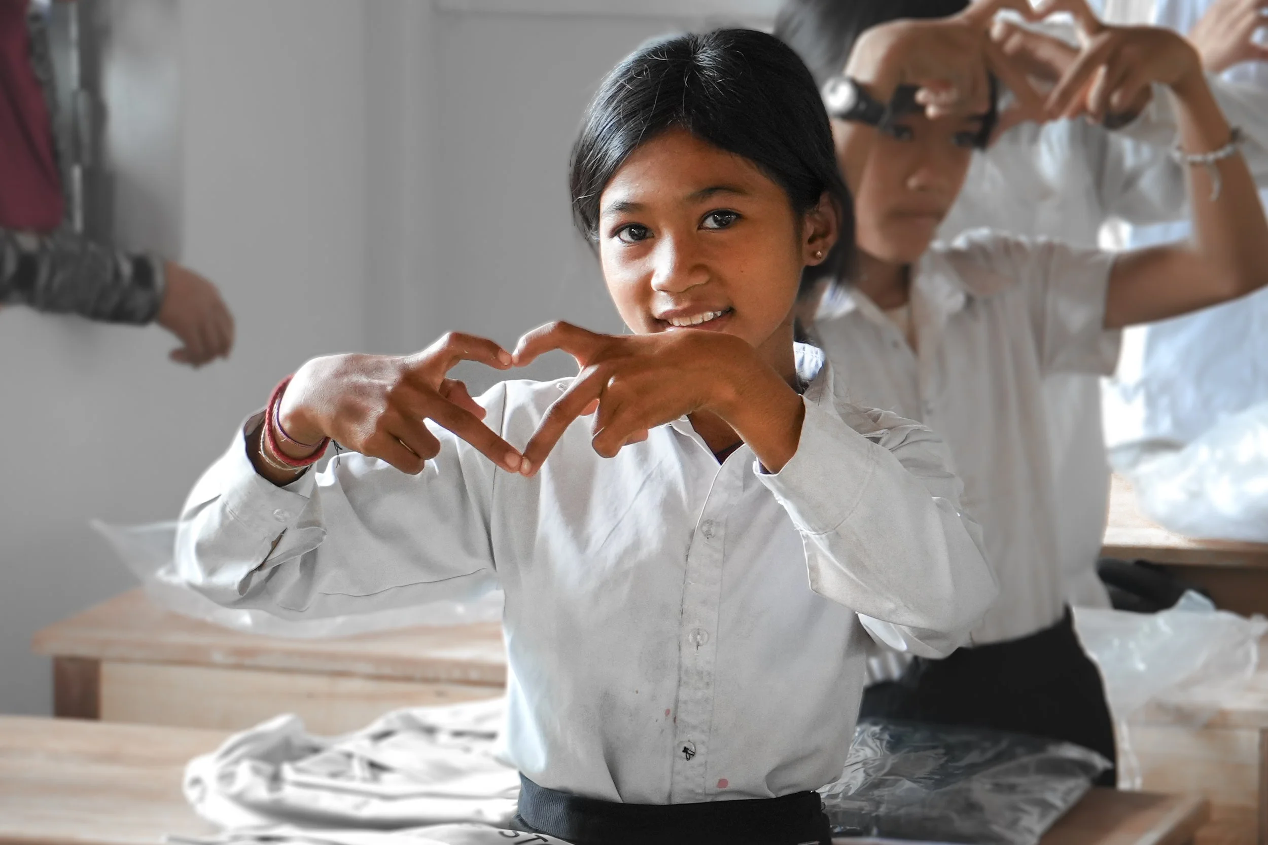 A young girl in a white shirt makes a heart shape with her hands in a classroom, with other children in white shirts behind her.