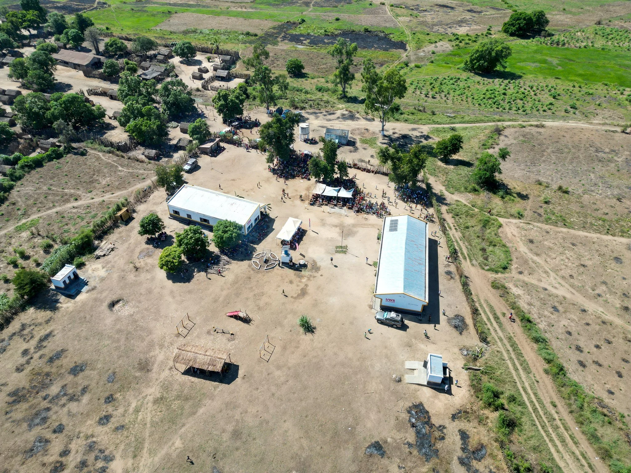 Aerial view of a rural area with small houses, trees, and open land. There is a gathering of people near a large building with a metal roof, some tents, and vehicles.