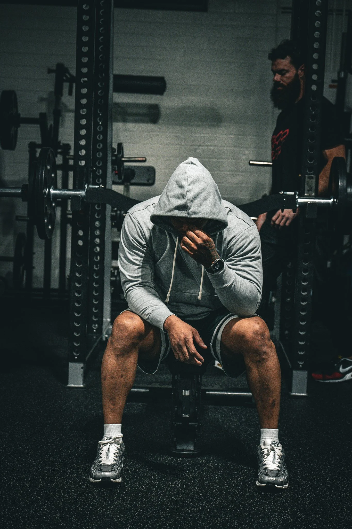 A man in a gray hoodie and shorts sitting on a bench in a gym, covering his face with his hand, with a hood over his head. Another man stands in the background near gym equipment.