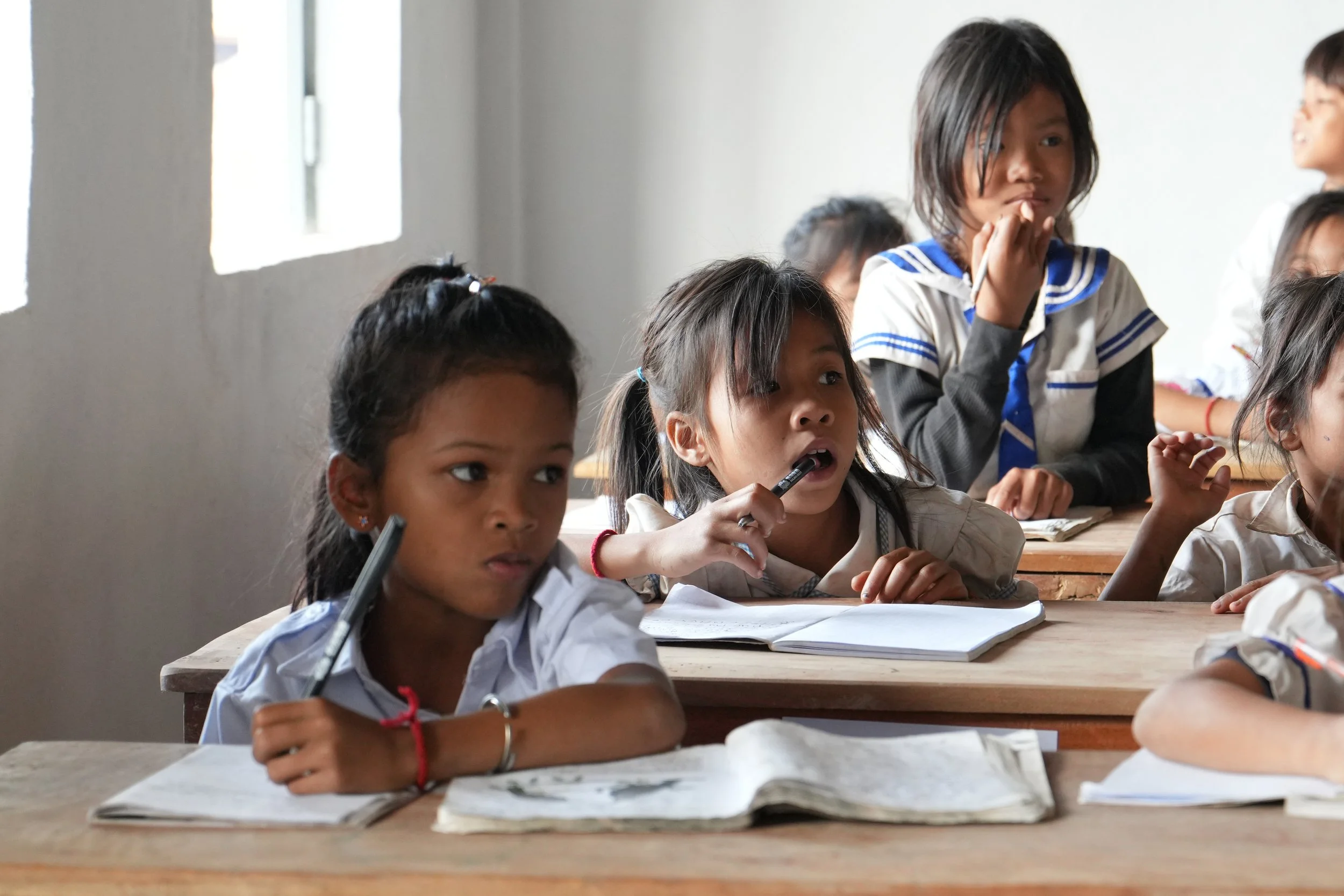Young students in a classroom, sitting at wooden desks, engaged in a lesson. Some have notebooks open, and one girl is holding a pen to her mouth.