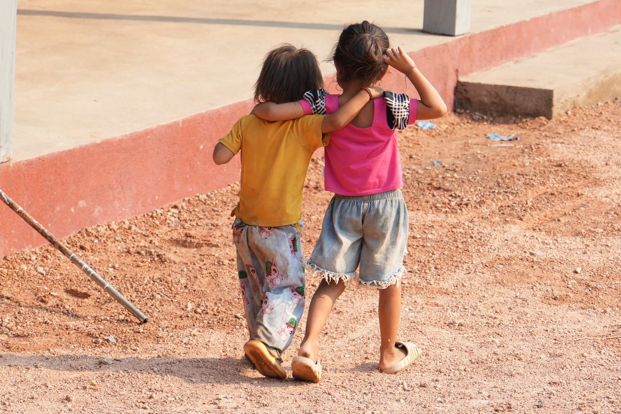 Two children, a boy in a yellow shirt and patterned pants and a girl in a pink top and shorts, walk together arm-in-arm on a dirt ground outside, with the girl resting her hand on her head.