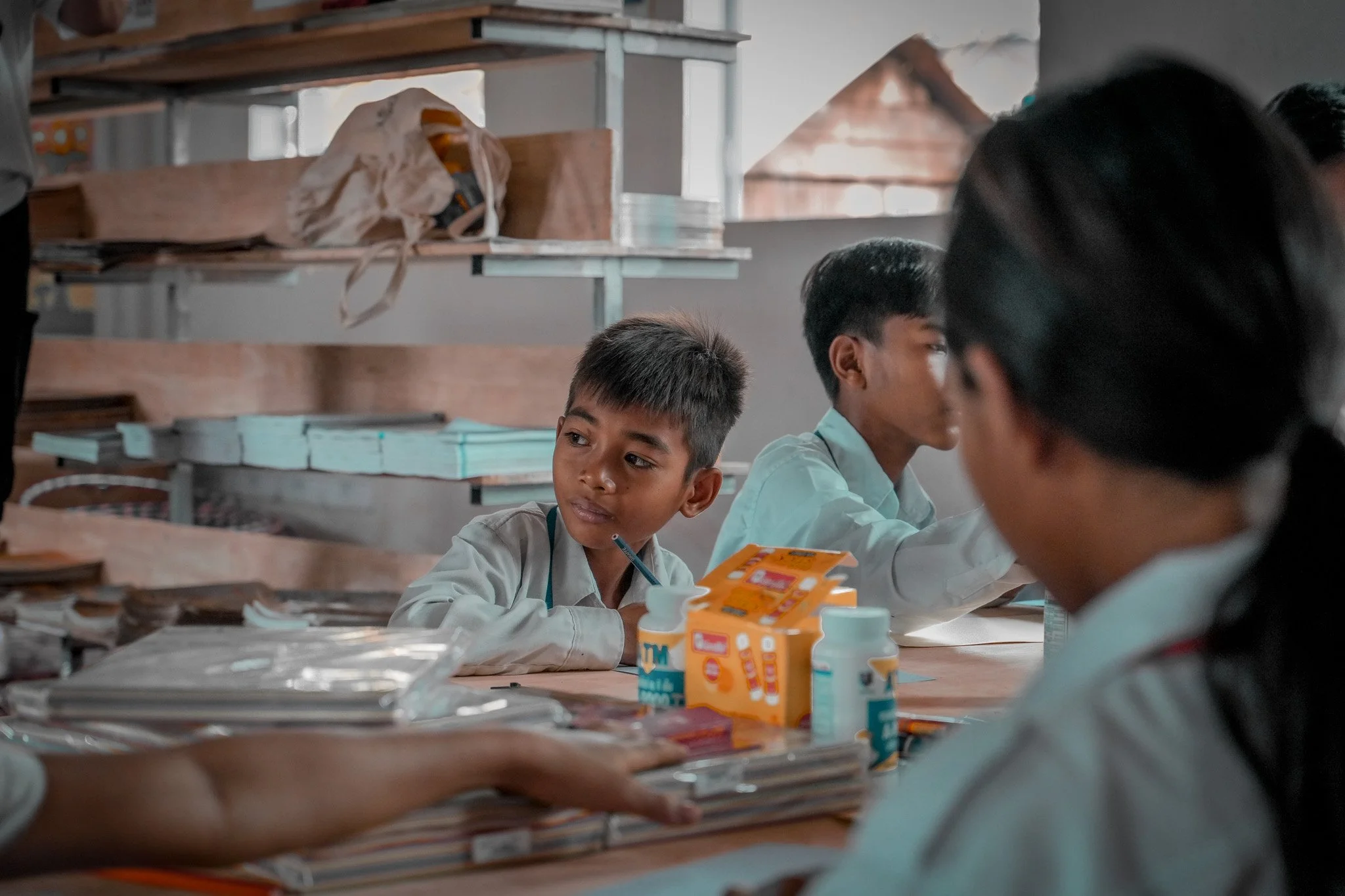 Children sitting at a table in a classroom, with books, notebooks, and medication bottles on the table, engaged in an activity or lesson.