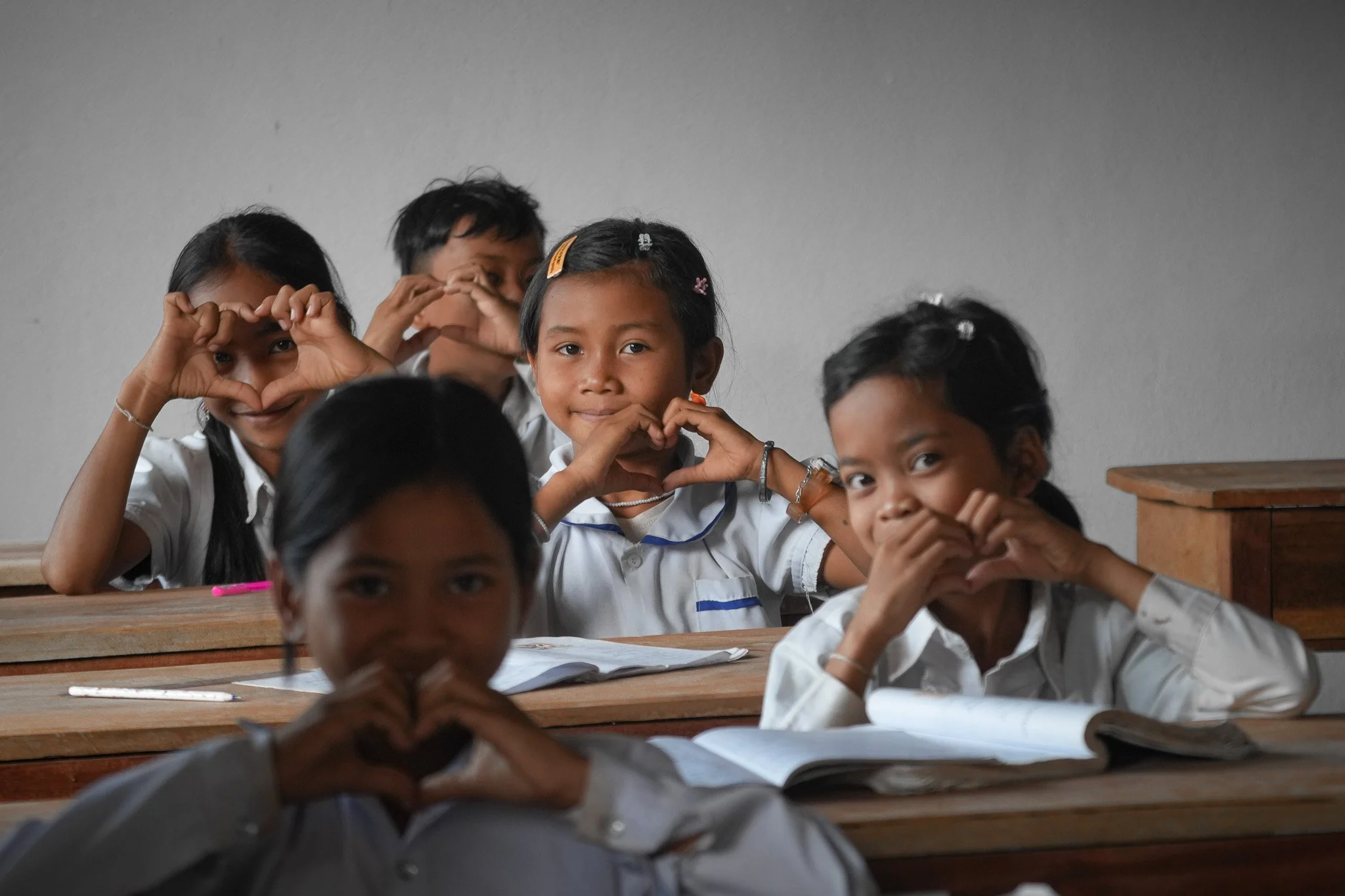 A group of five young students sitting at desks in a classroom, making heart shapes with their hands and smiling.