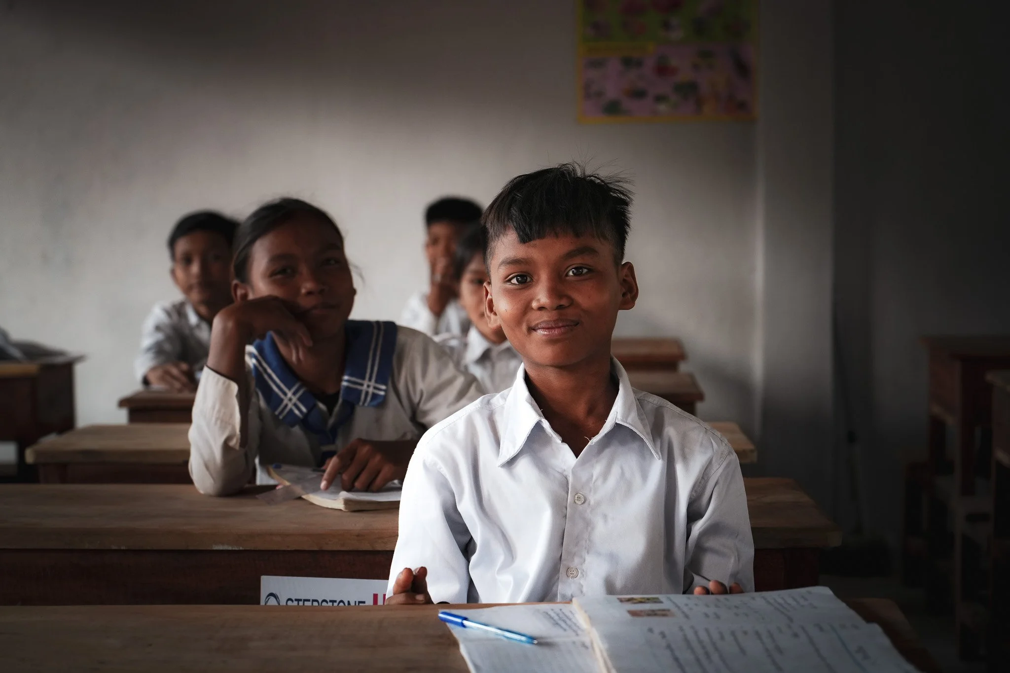 School children sitting at desks in classroom, smiling at camera.