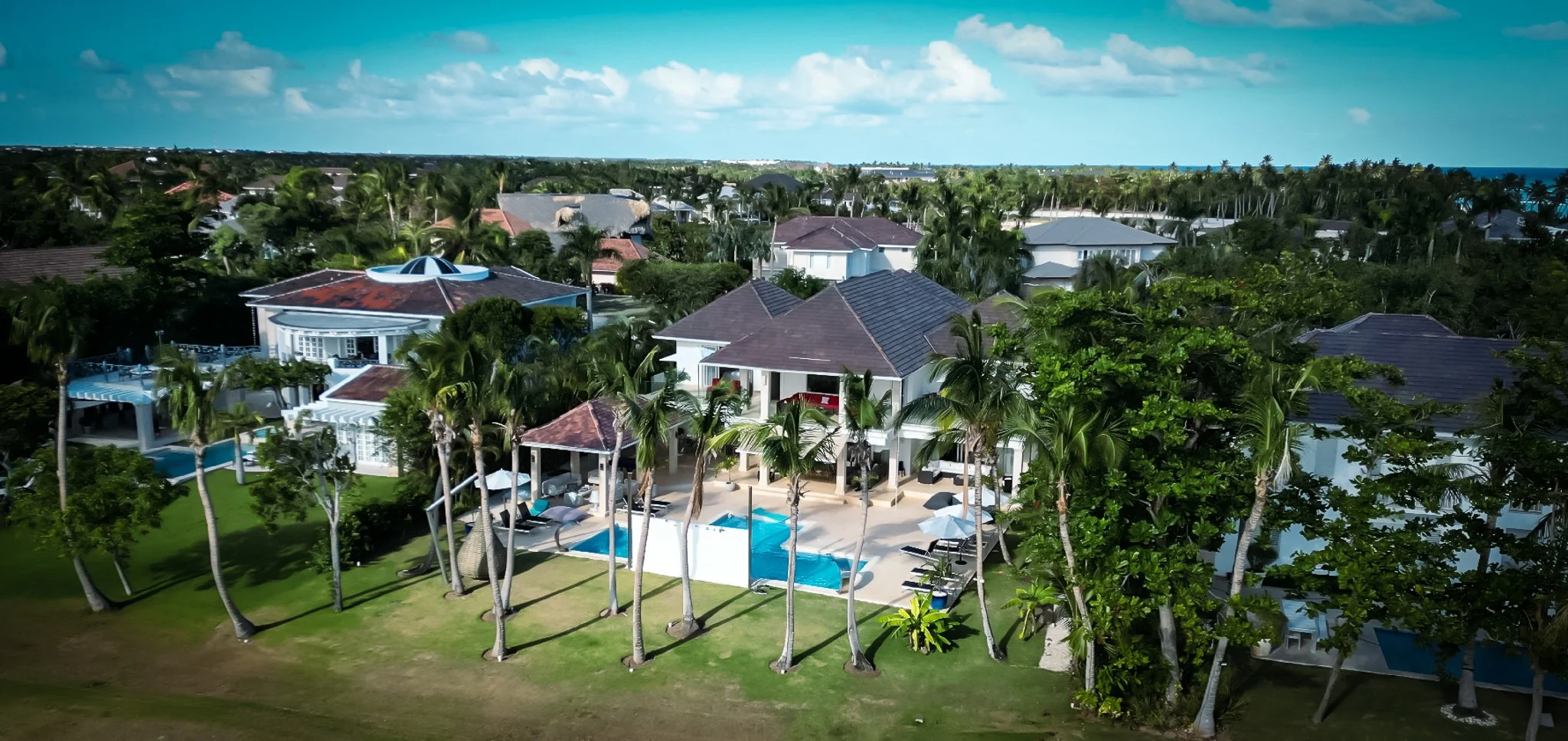 Aerial view of a seaside residential neighborhood with a large house featuring a swimming pool, surrounded by palm trees and other houses, on a sunny day.