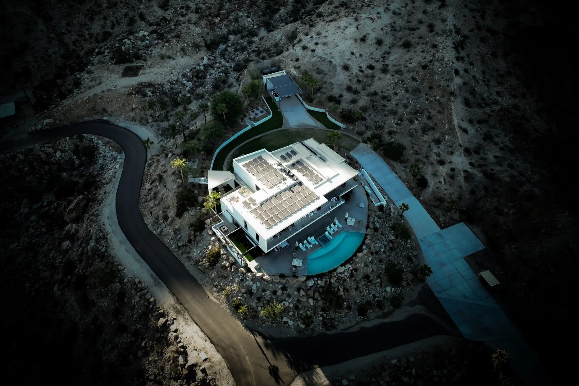 Aerial view of a modern house with solar panels on the roof, surrounded by a winding driveway, pool, and desert landscape.