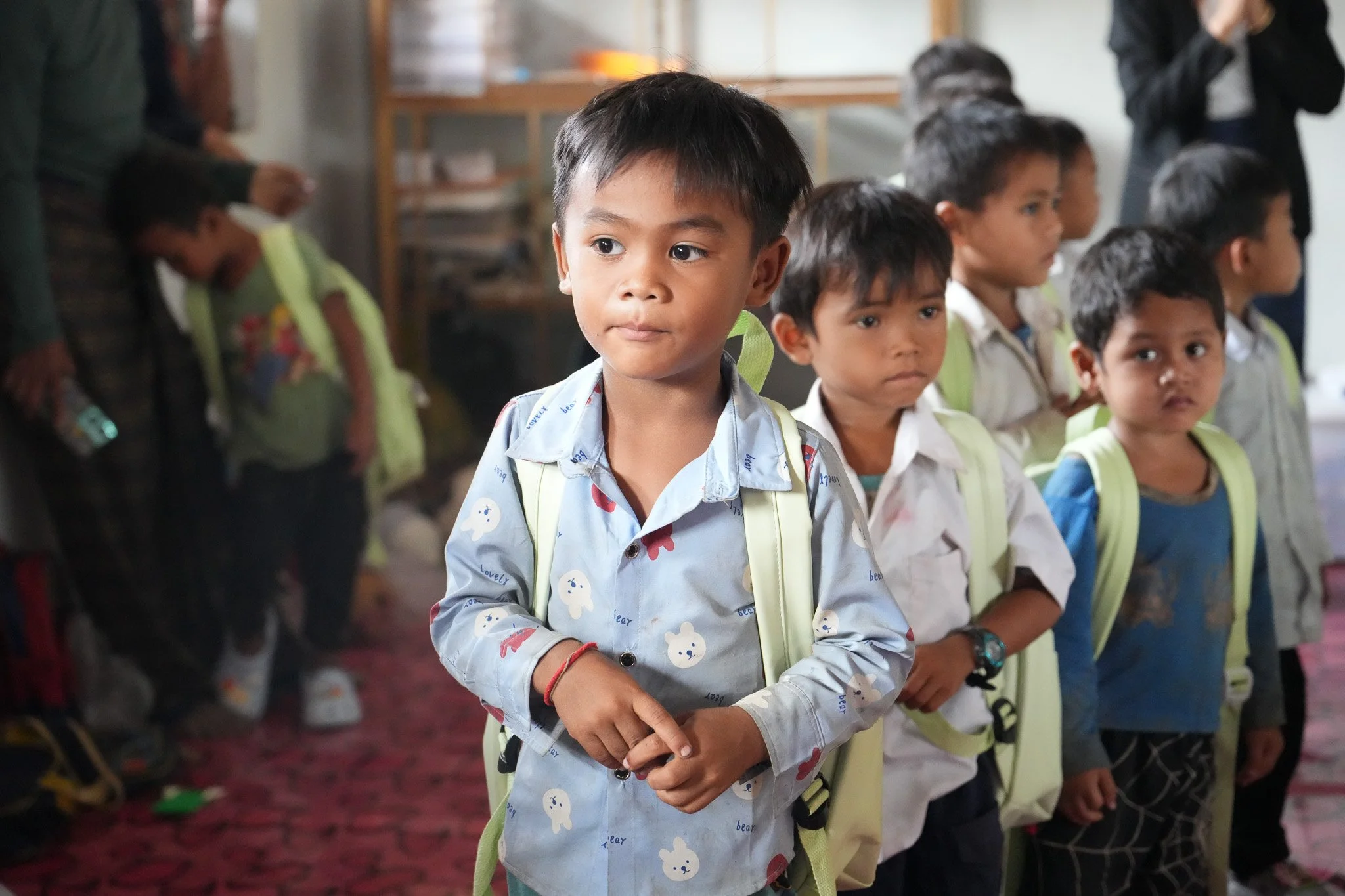 Young children, wearing school uniforms and carrying backpacks, lined up inside a classroom or school setting.