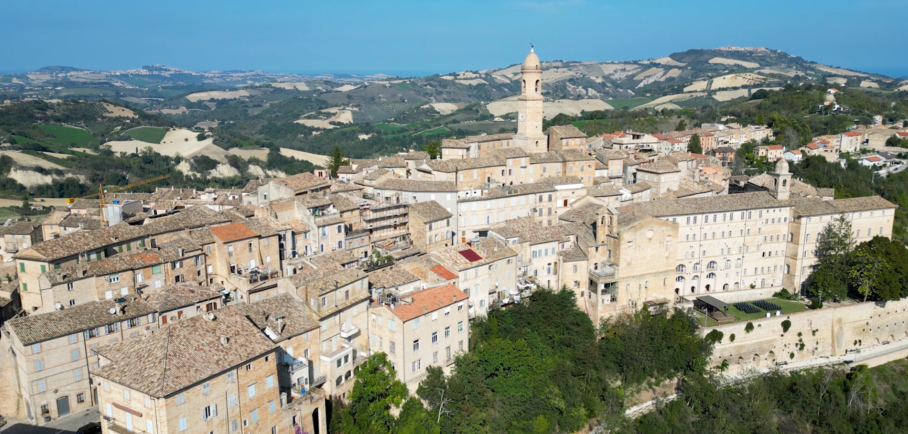 Aerial view of an old town with historic stone buildings, a tall clock tower, and surrounding rolling hills with patches of farmland and greenery.